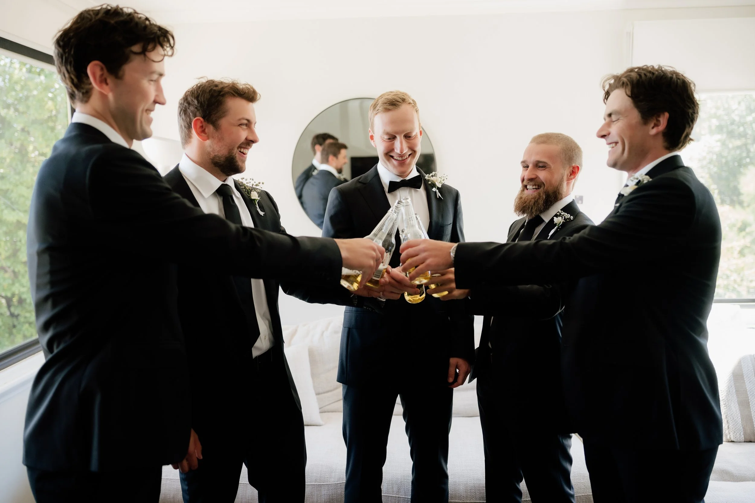 Five men in tuxedos raising beer bottles in a toast in a bright living room.