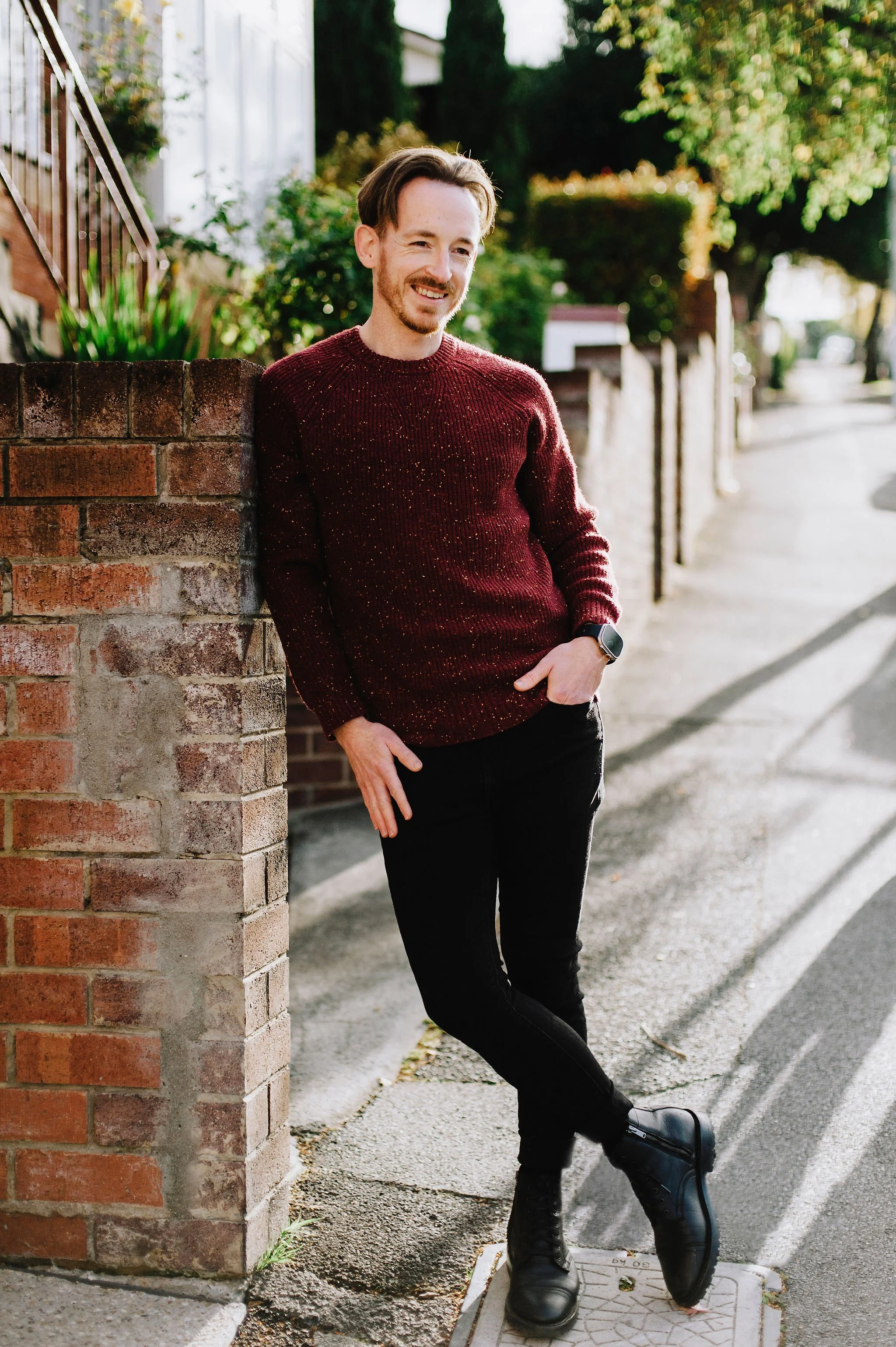 A man standing outdoors on a sidewalk, leaning against a brick wall, smiling, wearing a maroon sweater, black pants, black boots, and a smartwatch, with background of greenery and residential area.