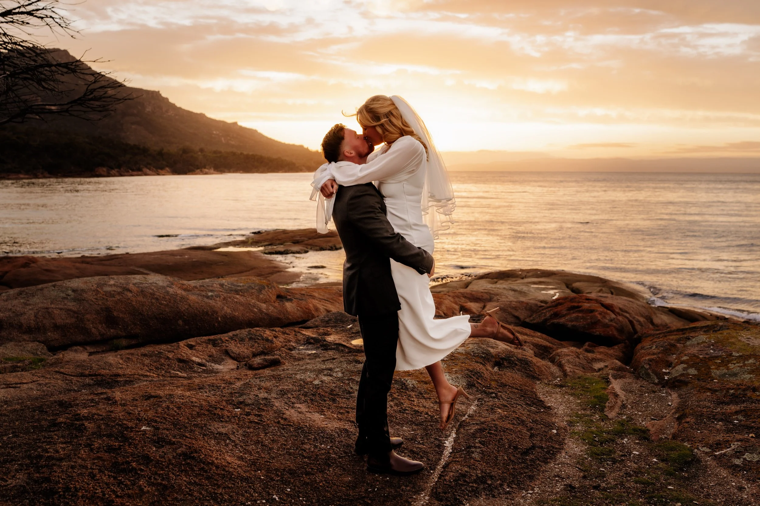 A bride and groom embrace on rocky shoreline at sunset, with the groom lifting the bride, who is wearing heels, against a backdrop of water, hills, and a cloudy sky.