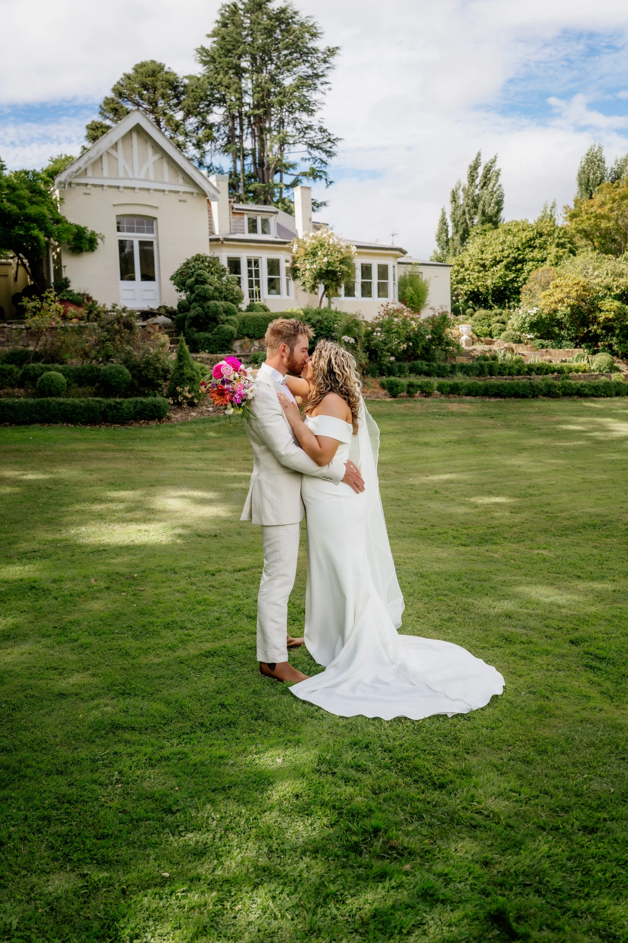 A bride and groom embracing on a lawn in front of a large white house surrounded by greenery and trees, during a wedding celebration.
