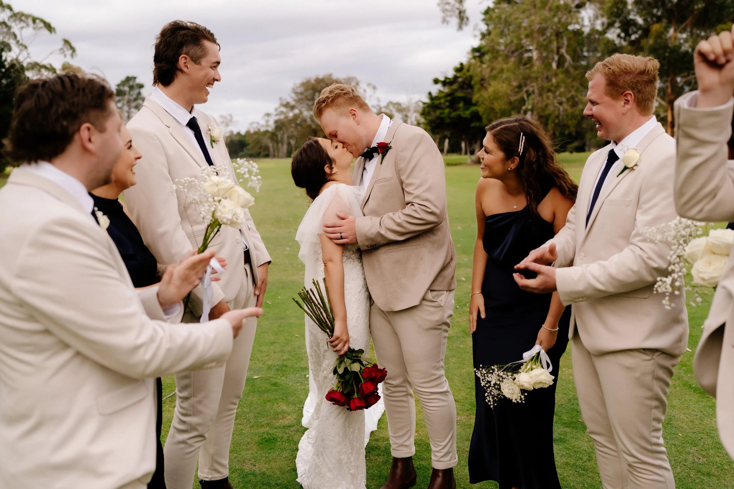 A wedding scene outdoors with a bride and groom kissing, surrounded by friends in formal attire, holding flowers, on a grassy area with trees in the background.