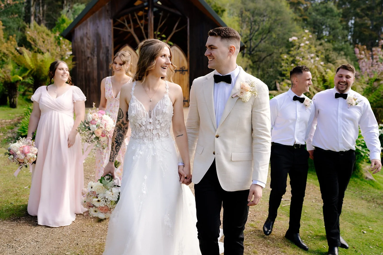 A wedding party walking outdoors with the bride and groom in front, holding hands and smiling at each other, surrounded by bridesmaids and groomsmen, with a wooden shed and lush green trees in the background.