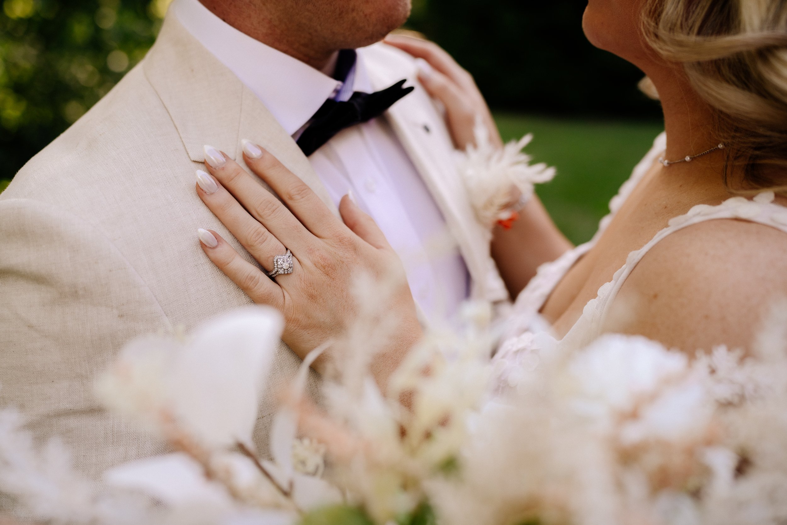 Close-up of a couple at their wedding, showing a woman's hand with a diamond ring on her finger resting on a man's beige suit jacket. The man is wearing a white shirt and black bow tie, and the woman is wearing a white dress with floral details and a