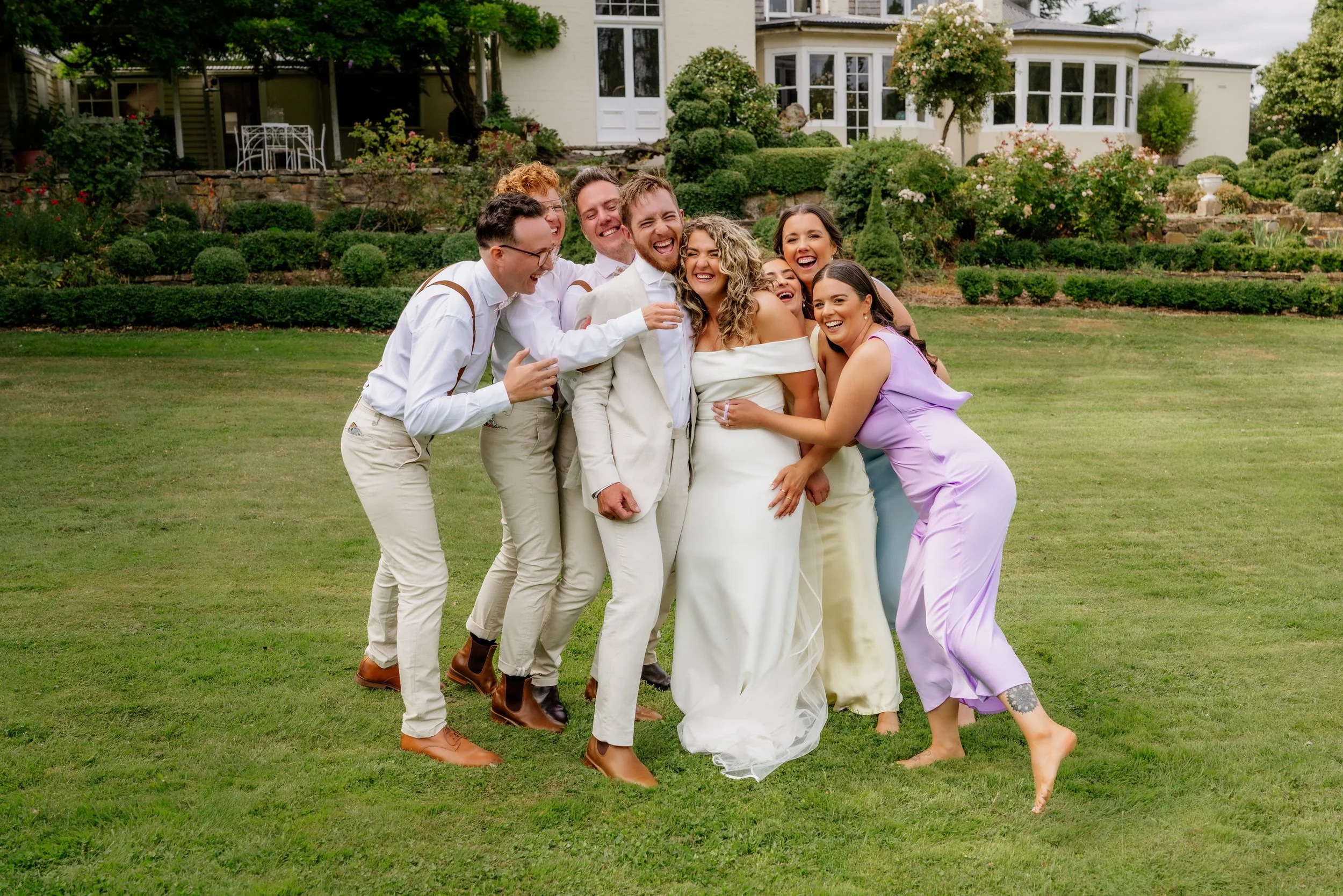 Group of people in wedding attire laughing and hugging on a lawn