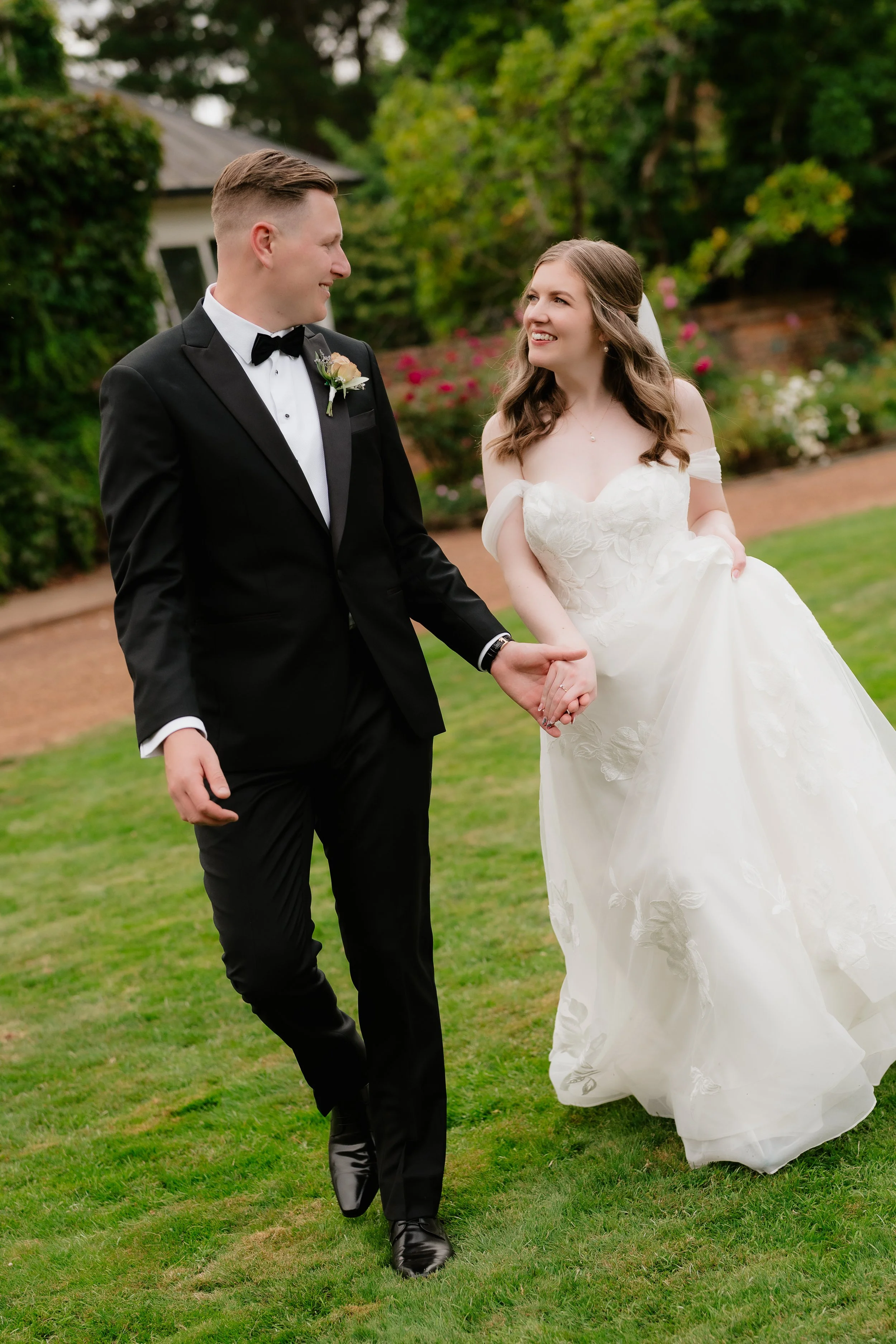 A bride and groom walking hand in hand outdoors on a wedding day, smiling at each other, with greenery and flowers in the background.