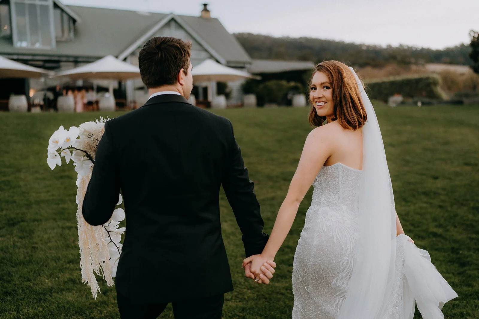 A bride and groom holding hands outdoors at a wedding venue, with the bride smiling and looking back. The bride is in a strapless white wedding gown with a veil, and the groom in a black suit. The background features a grassy area and a building with
