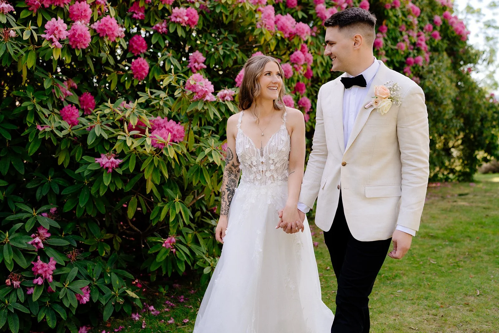 A bride and groom holding hands outdoors with pink flowering bushes in the background. The bride wears a lace wedding dress and has tattoos on her left arm. The groom wears a white tuxedo jacket, black pants, and a black bow tie.