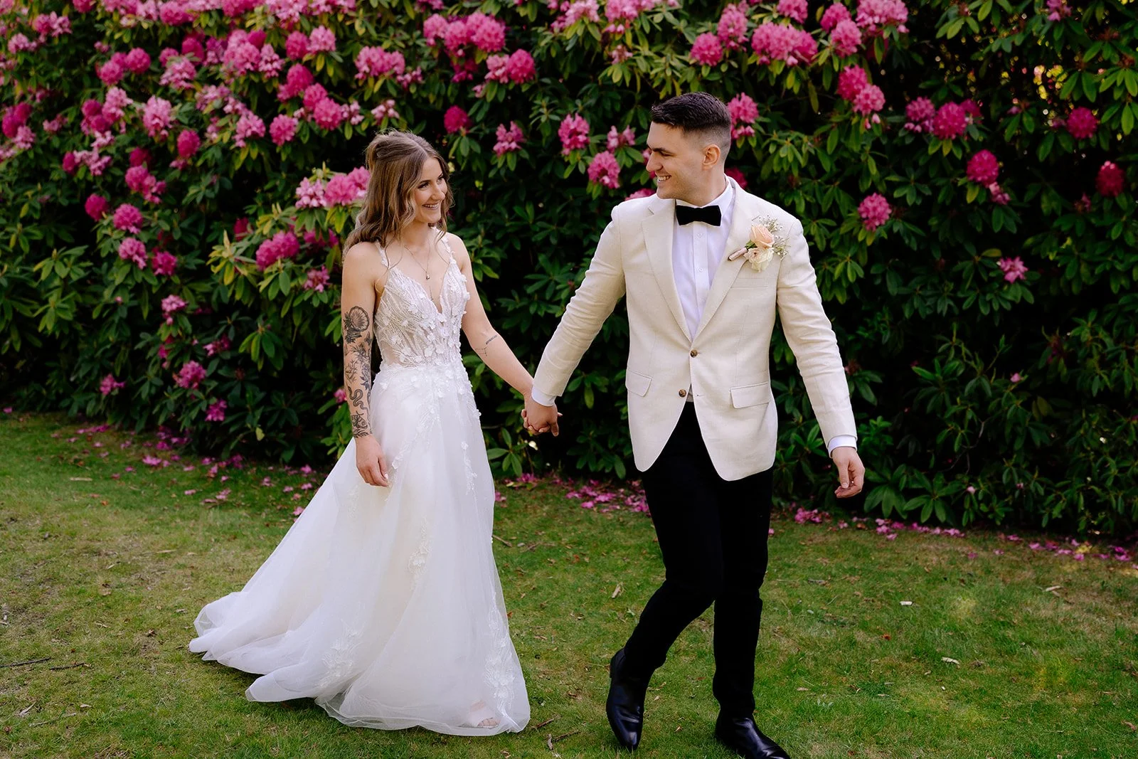 A newlywed couple holding hands and smiling at each other outdoors. The bride wears a white wedding gown with lace details and has tattoos on her arms. The groom is dressed in a white tuxedo jacket, black trousers, and a black bow tie, with a boutonn