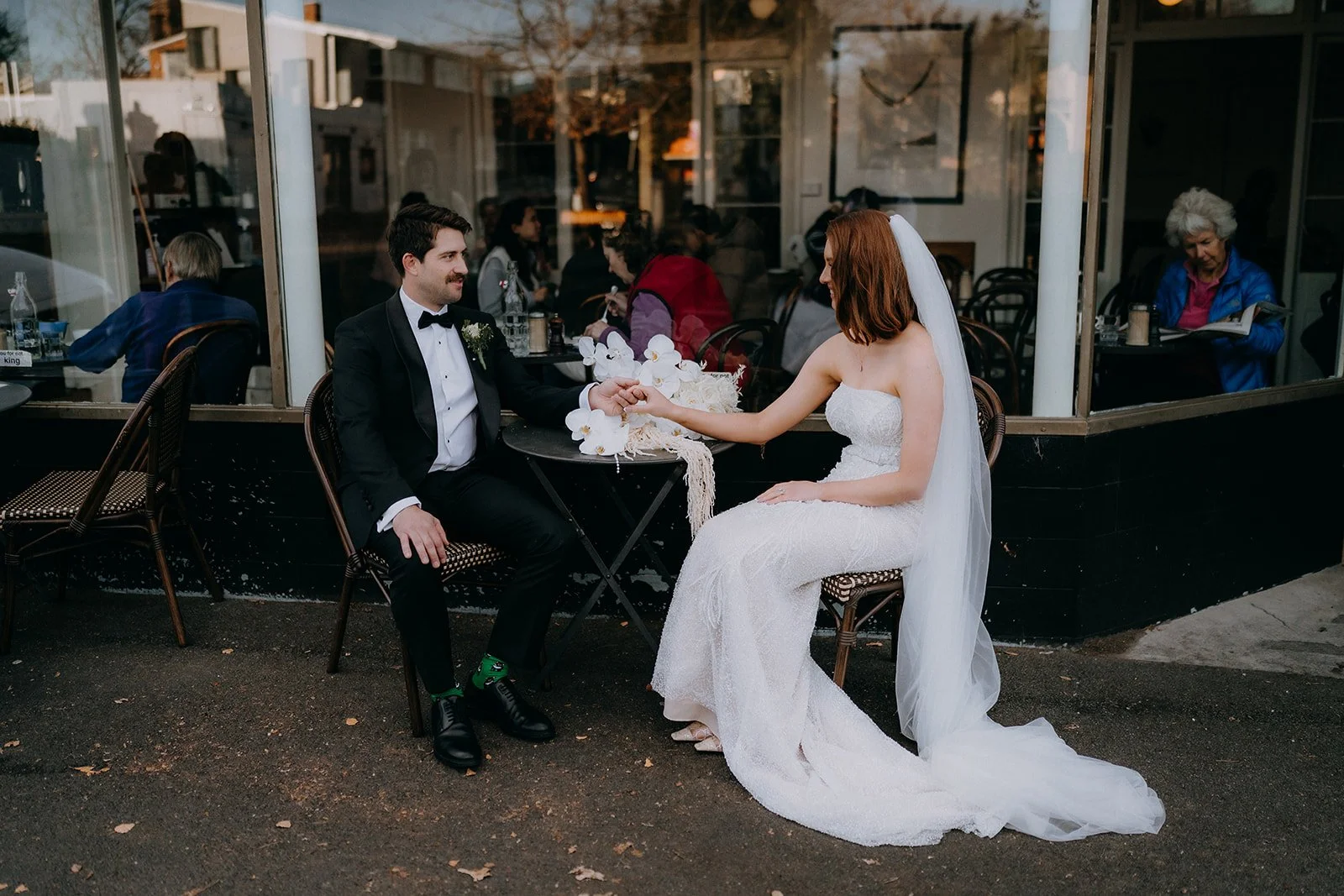 A bride and groom sitting outside a cafe during their wedding, holding hands across a small table with white flowers, with guests visible inside through the window.