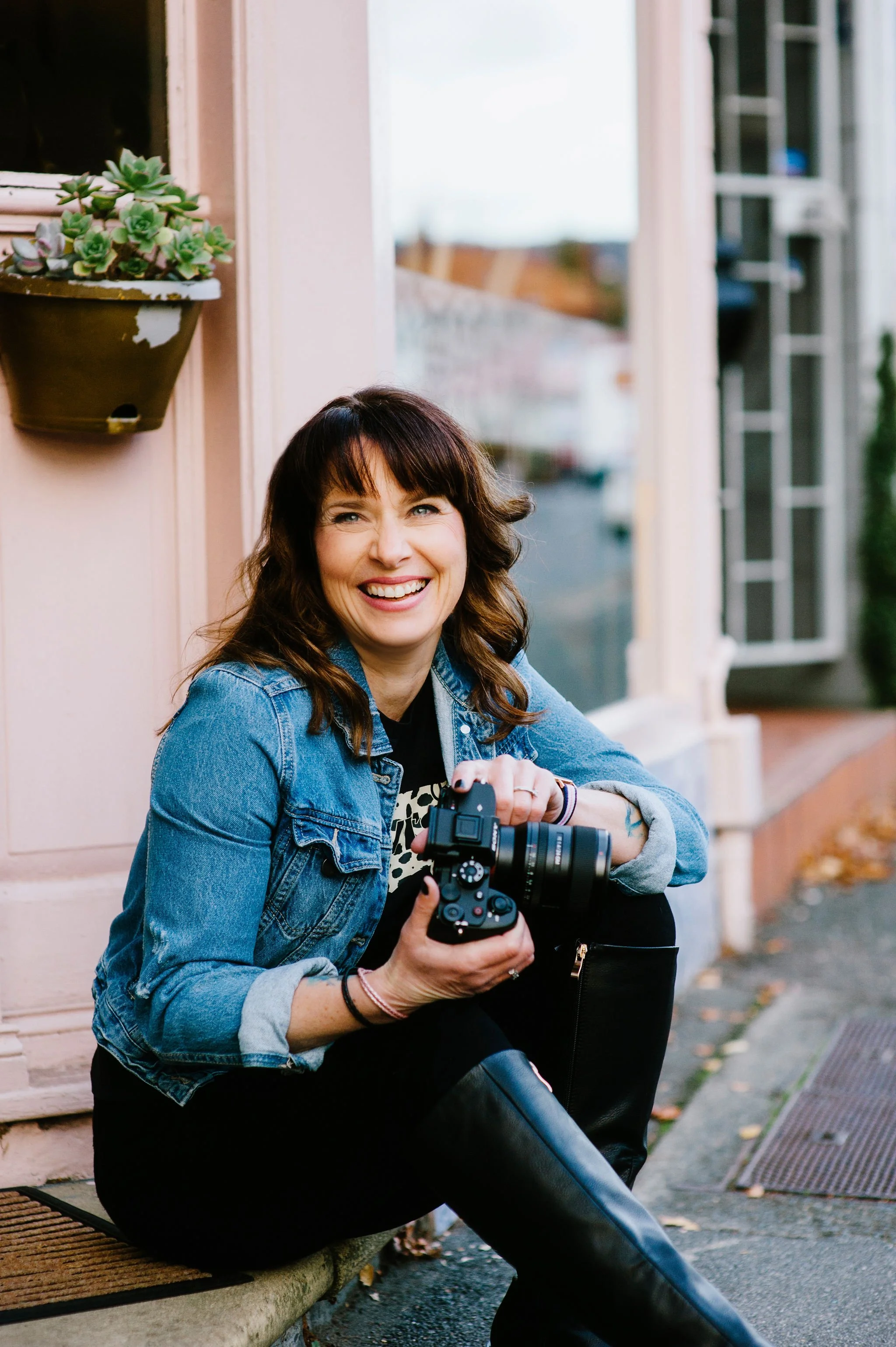 A woman with brown hair sitting outside on a sidewalk, smiling, holding a camera, wearing a denim jacket and black pants.
