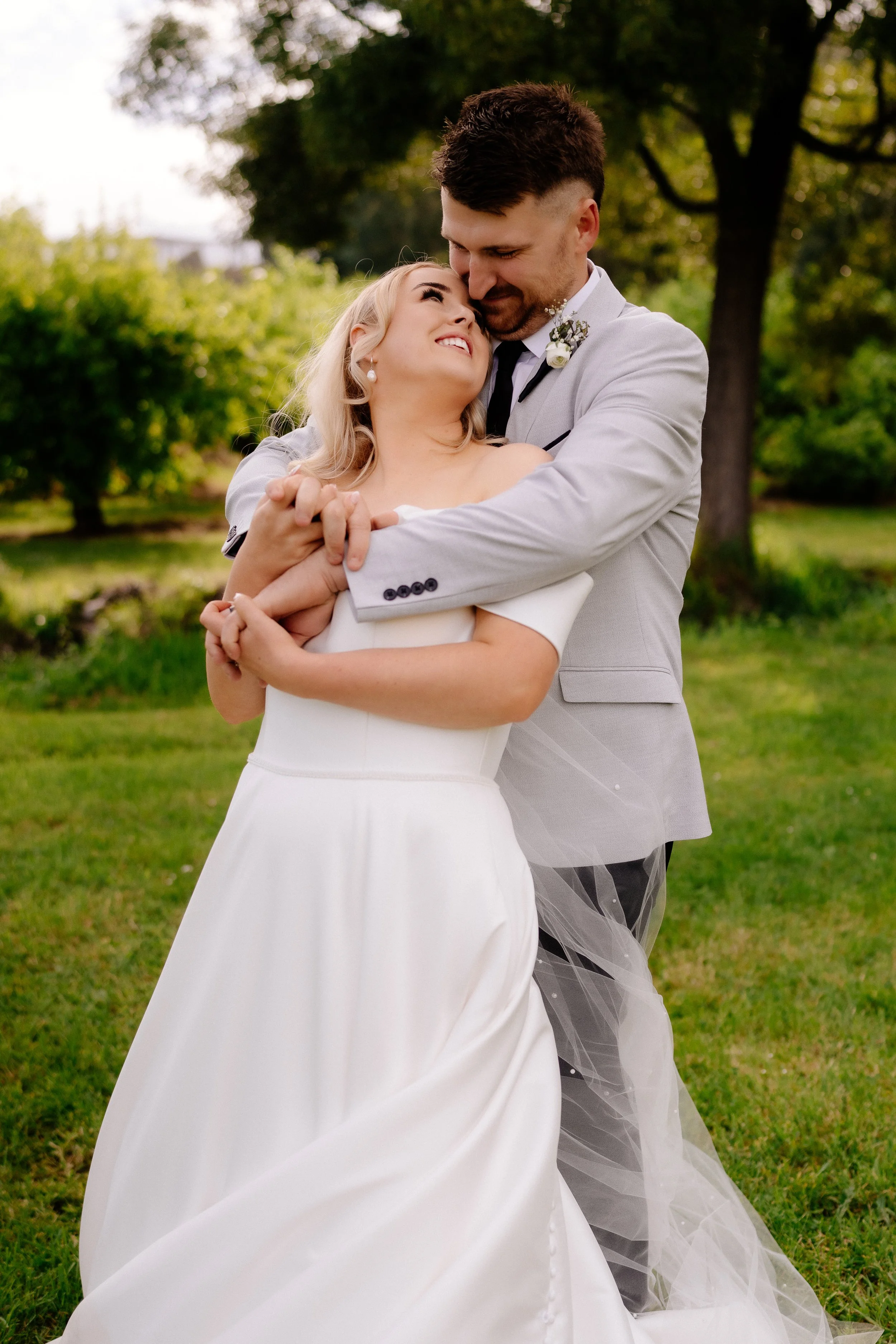 A bride and groom embracing outdoors on their wedding day, with trees and greenery in the background.