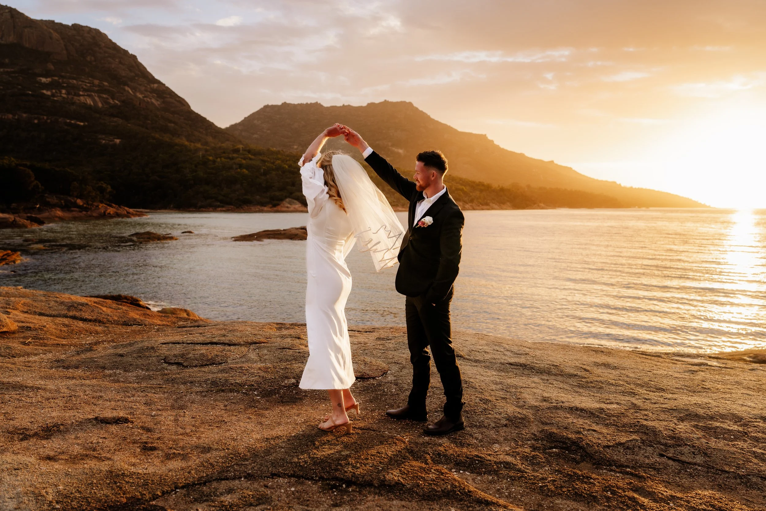 A bride and groom dancing on a rocky shoreline during sunset, with mountains and water in the background.