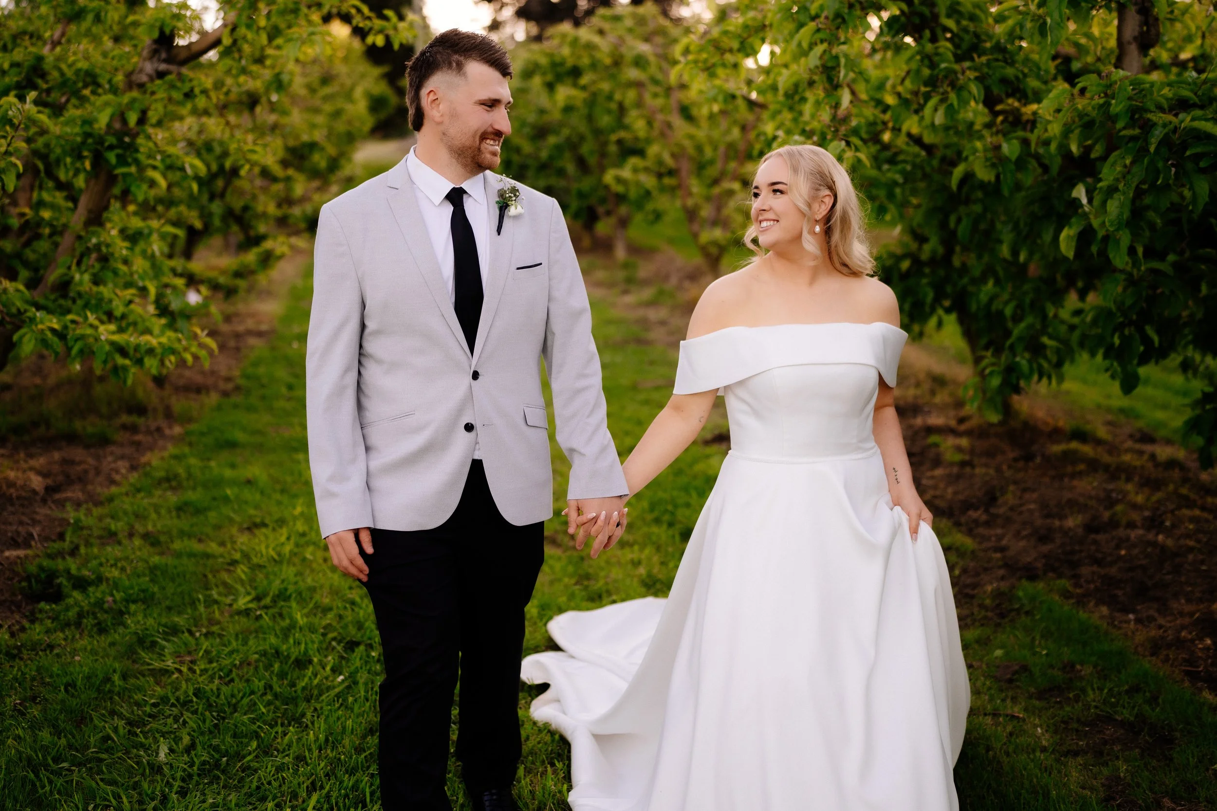 A bride and groom holding hands and smiling at each other in an orchard with green trees.