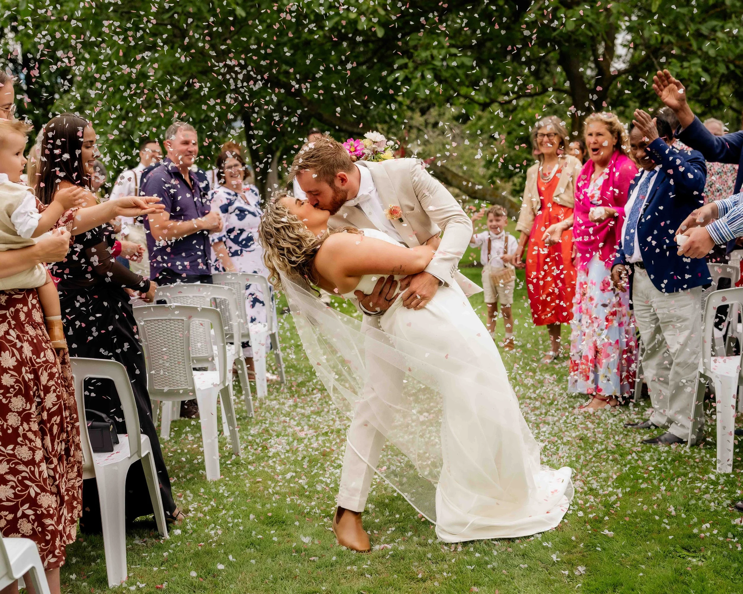A bride and groom share a kiss while leaning back in a dip during their outdoor wedding ceremony, surrounded by guests throwing confetti on a grassy field with trees.