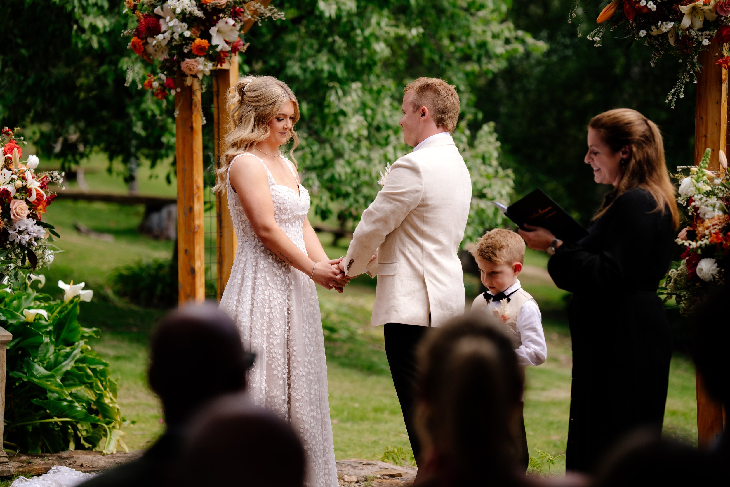 Couple exchanging wedding vows during an outdoor ceremony, with an officiant, a young boy in a tuxedo, and guests in the background.