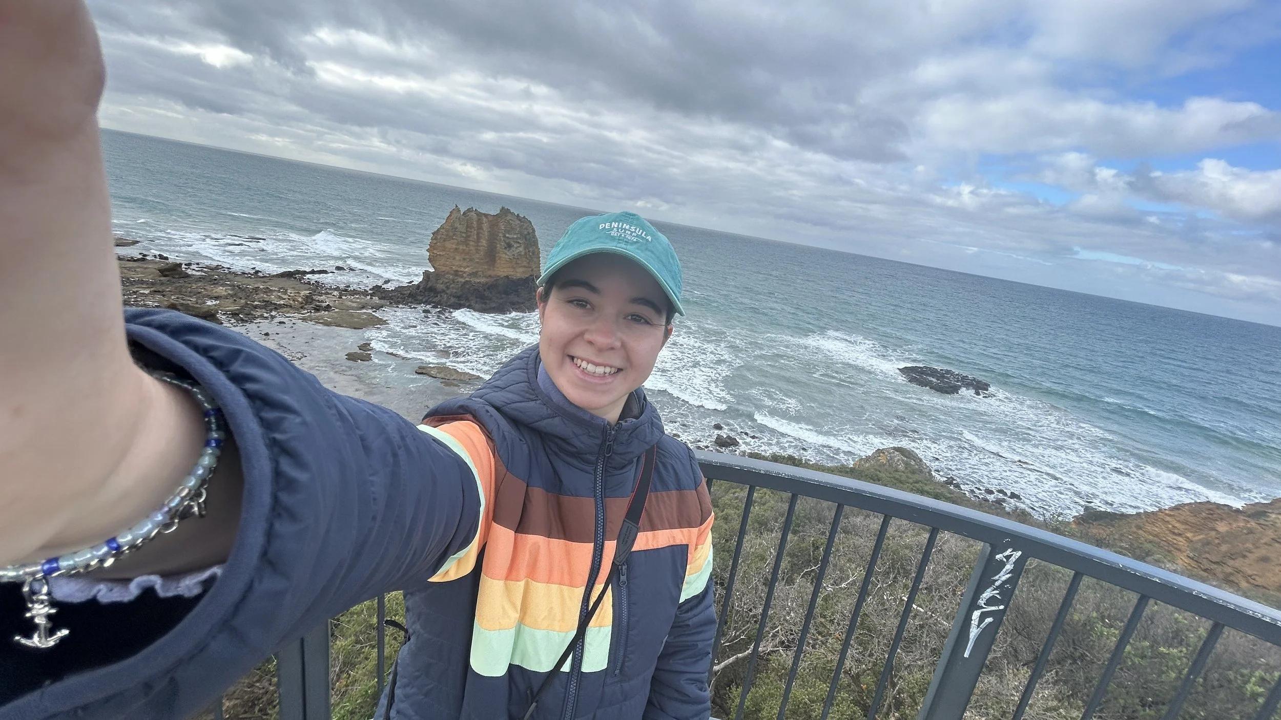 A young woman smiling and taking a selfie at a seaside viewpoint, with rugged rocks and the ocean in the background.