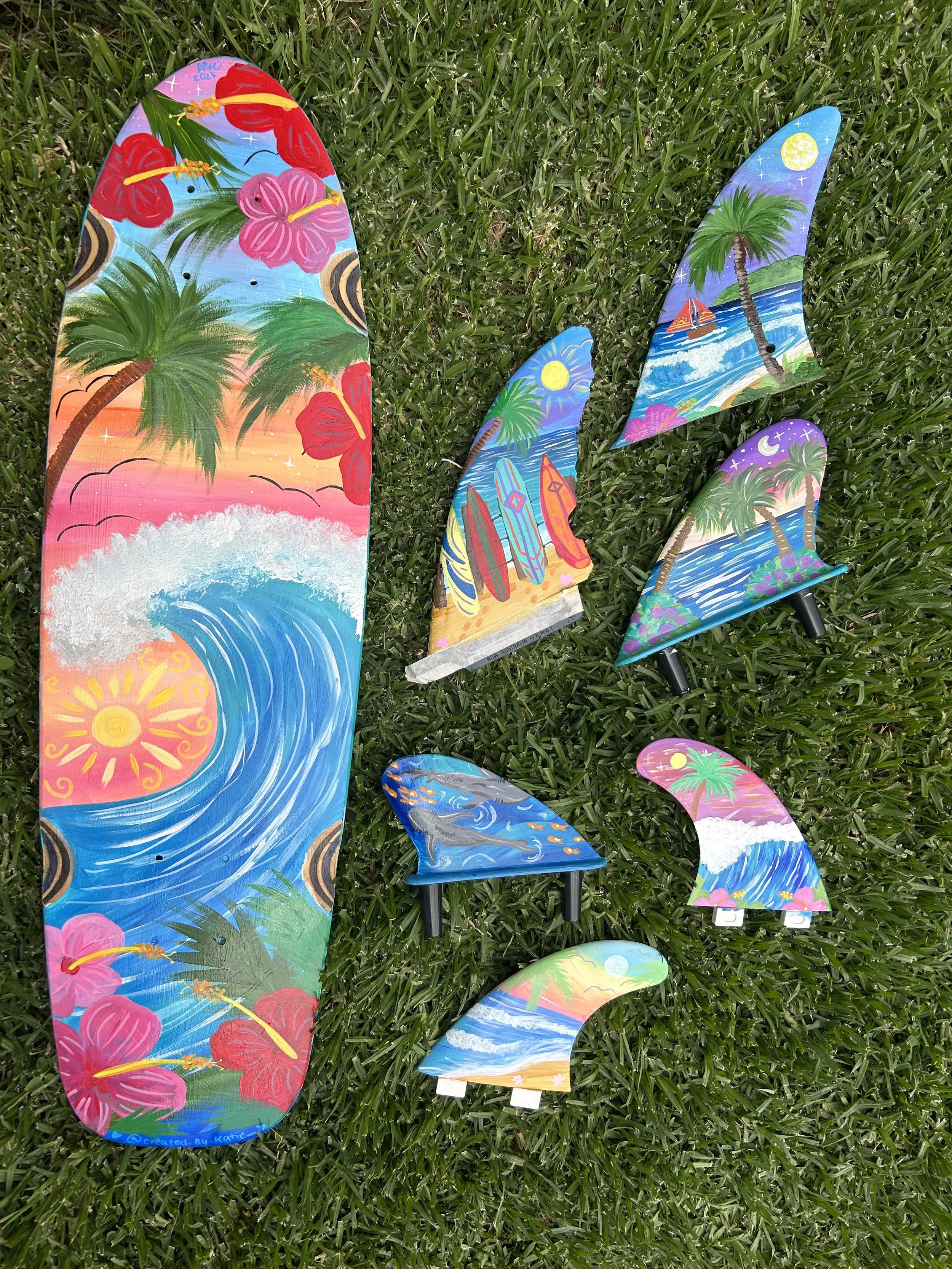 A colorful collection of painted surfboard and skateboard fins spread on grass, featuring tropical and ocean-themed designs with waves, palm trees, sun, and moon.