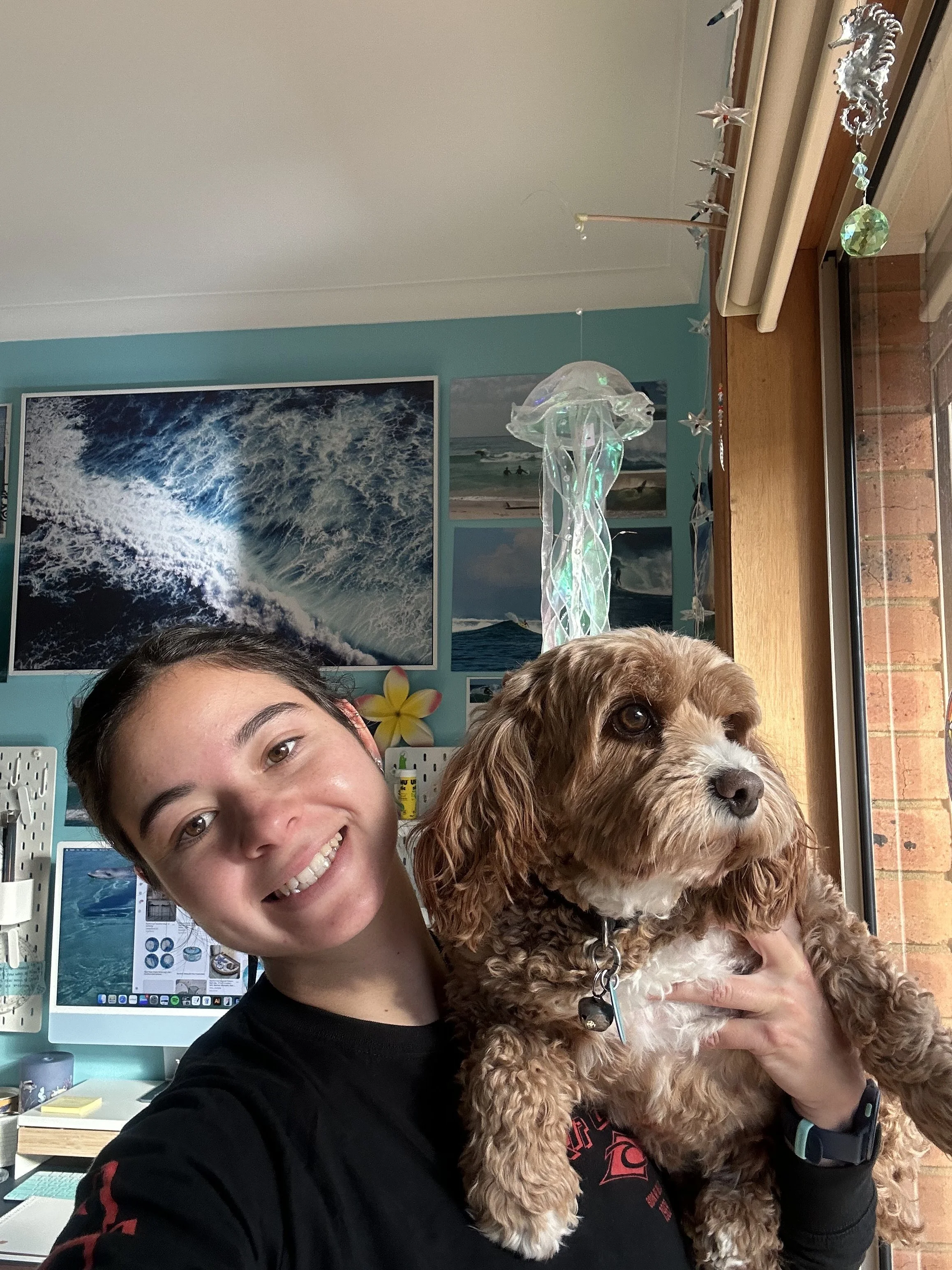 A young woman smiling and holding a brown and white curly-haired dog inside a room decorated with ocean-themed pictures on the wall and a decorative hanging lamp.