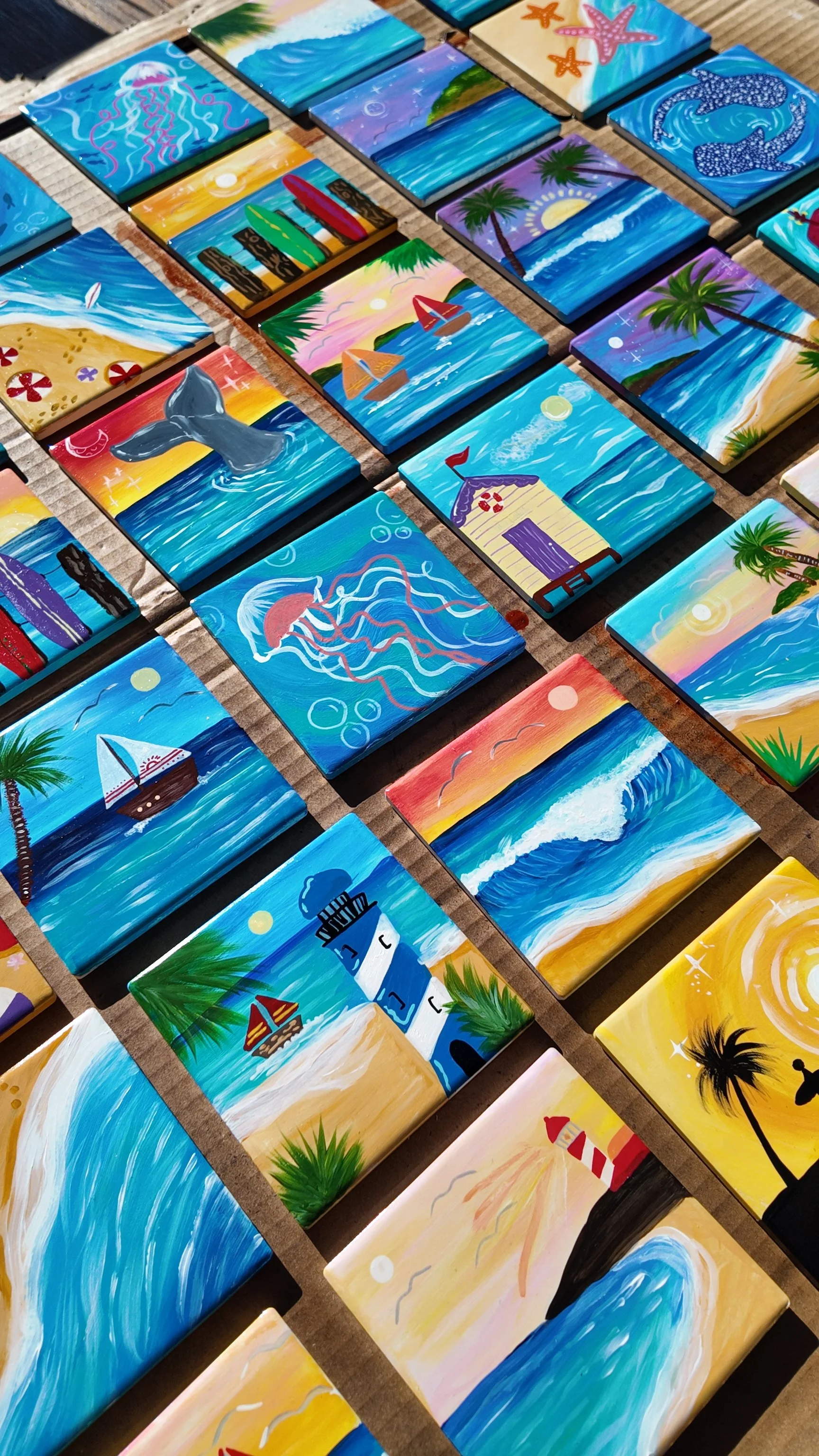 Collection of small, colorful, hand-painted tiles featuring beach and ocean scenes, including waves, lighthouses, sailboats, whales, sunsets, palm trees, and marine life.