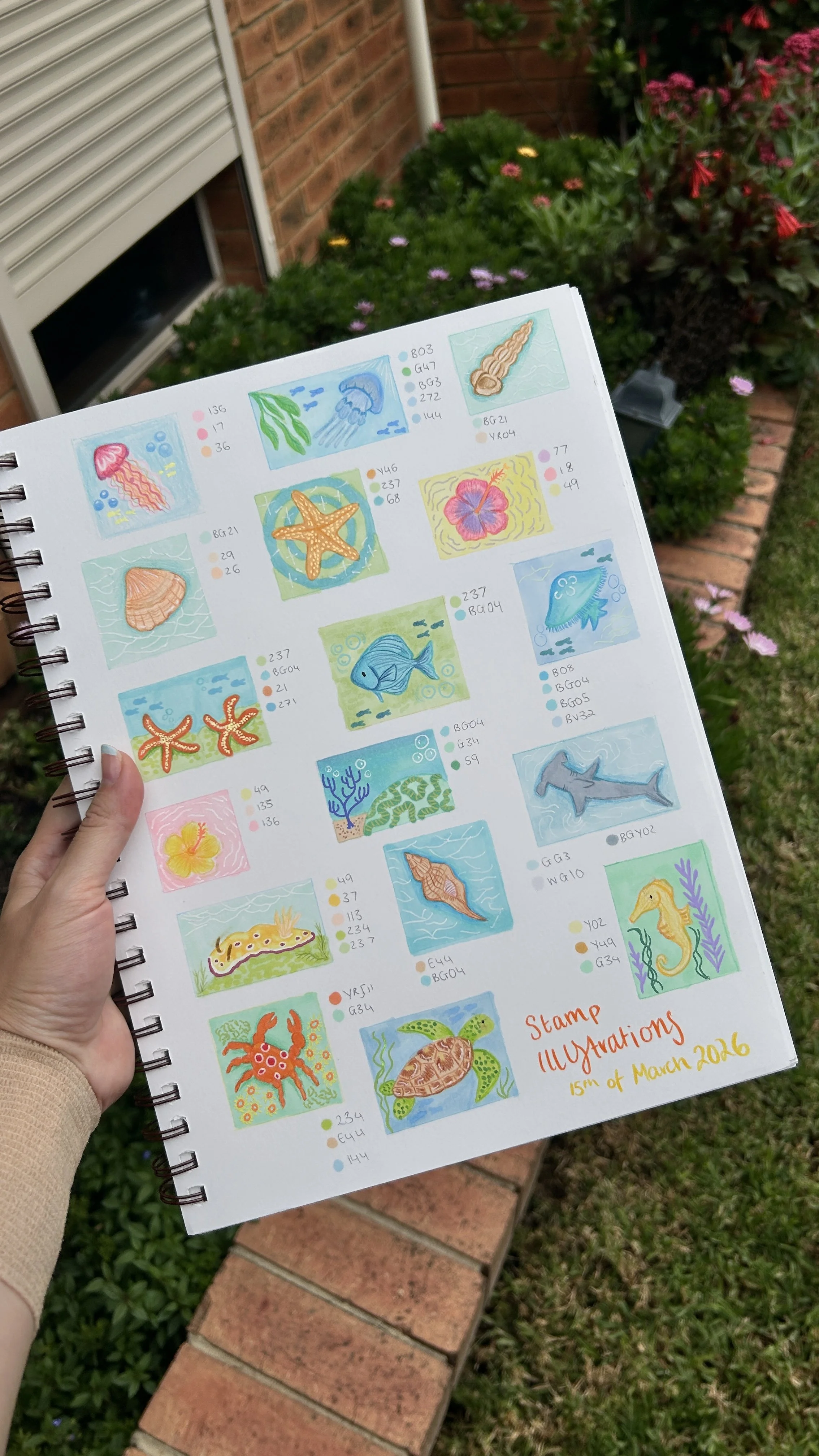 A hand holding a colorful illustrated booklet page with various underwater sea creatures and coral, each with color swatches and codes, titled 'Stamp Illustrations 15th of March 2016,' set outdoors against a garden with brick pathway and pink flowers