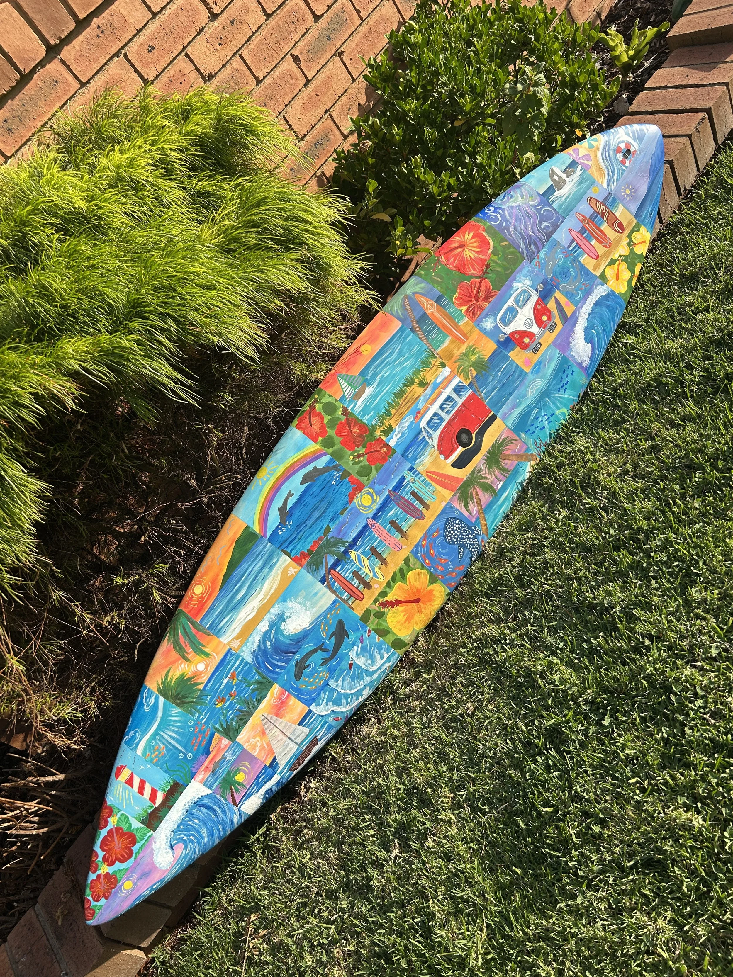 Colorful surfboard decorated with tropical, beach, and ocean-themed artwork, resting on grass beside a brick wall and lush green plants.