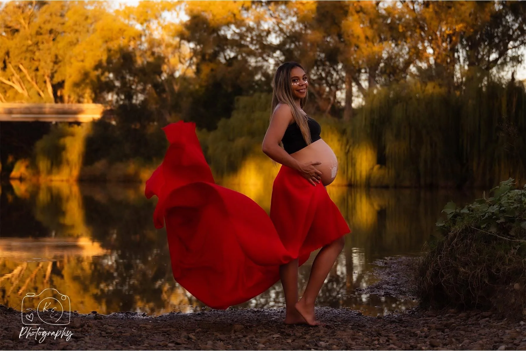 A pregnant woman standing barefoot by a river during autumn, wearing a black top and a flowing red skirt, smiling and touching her belly.