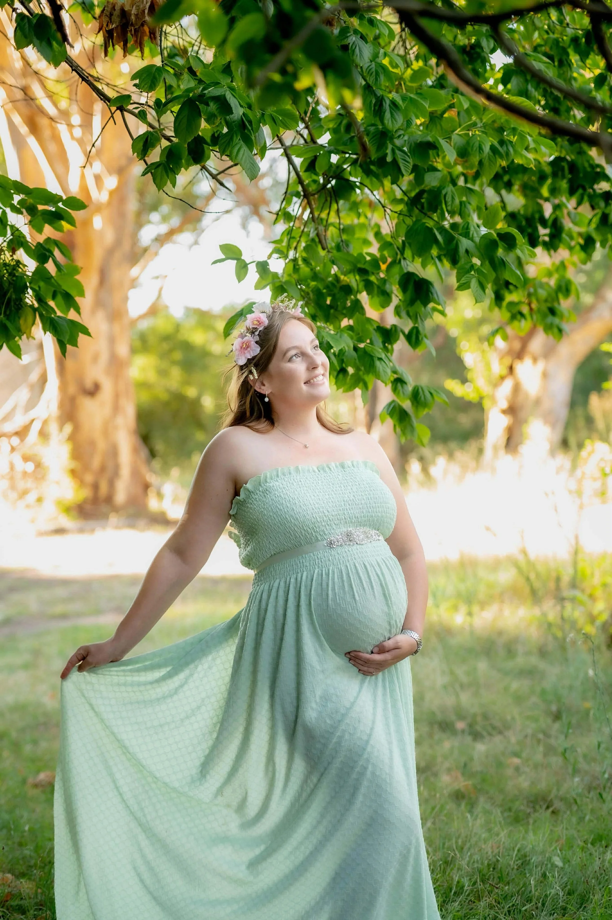 Pregnant woman in a light green dress with a floral headband outdoors, standing under a tree with sunlight filtering through the leaves.