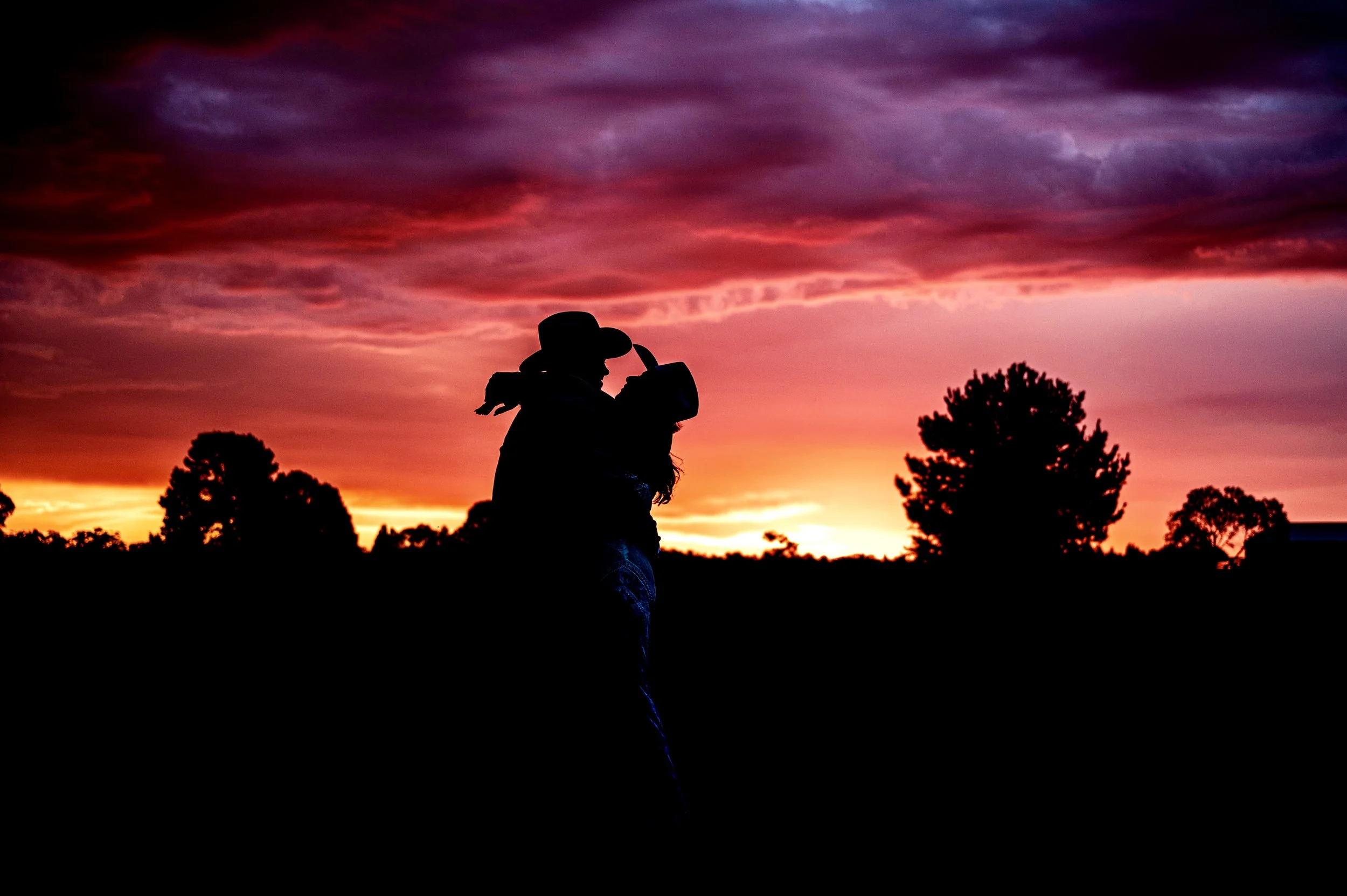 Silhouette of a person wearing a cowboy hat and holding a camera during a vibrant sunset with colorful sky and trees.