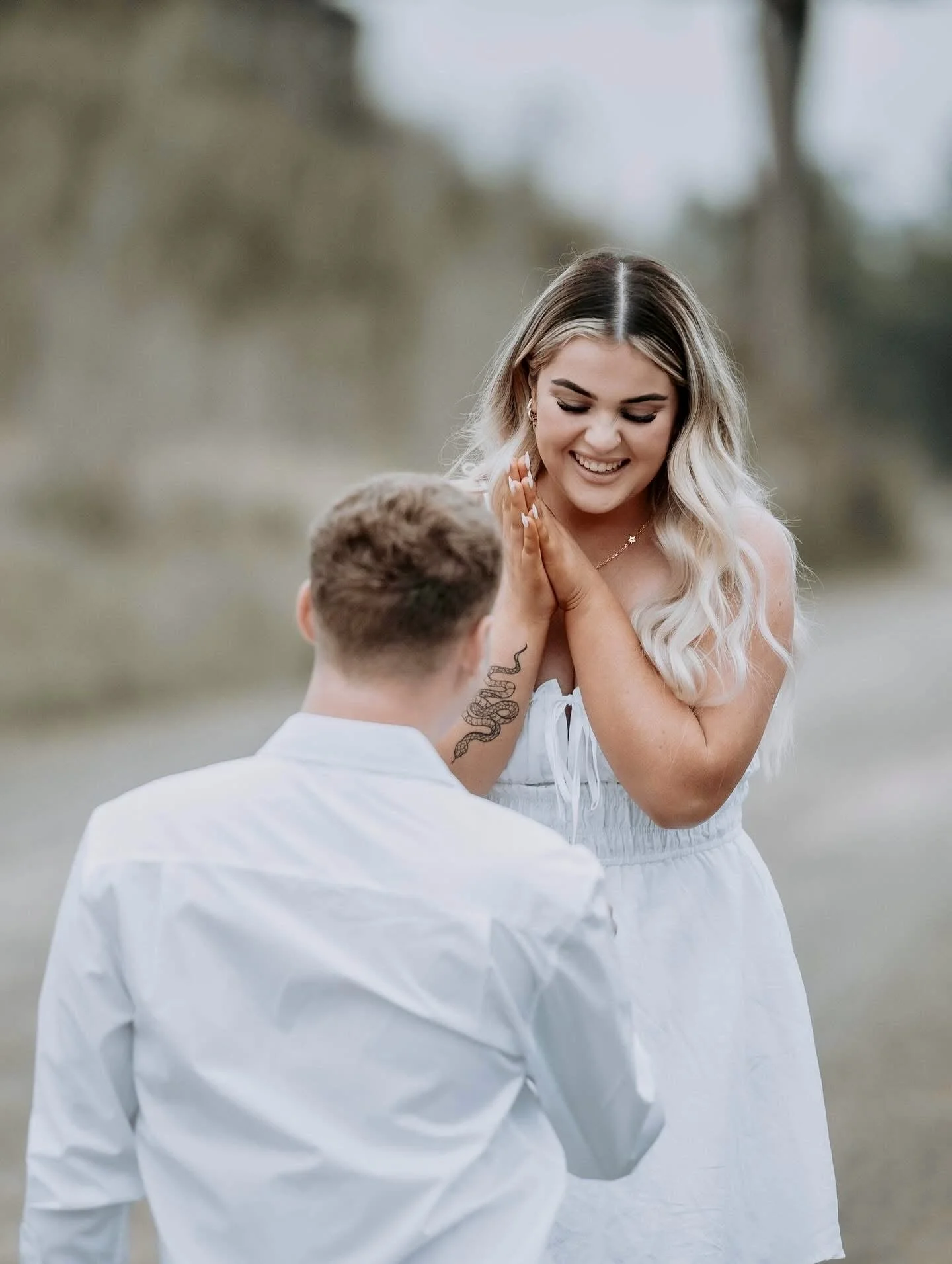 A woman and a man are engaged in a proposal outdoors. The woman is smiling with her hands clasped together, and the man is kneeling with his back to the camera.