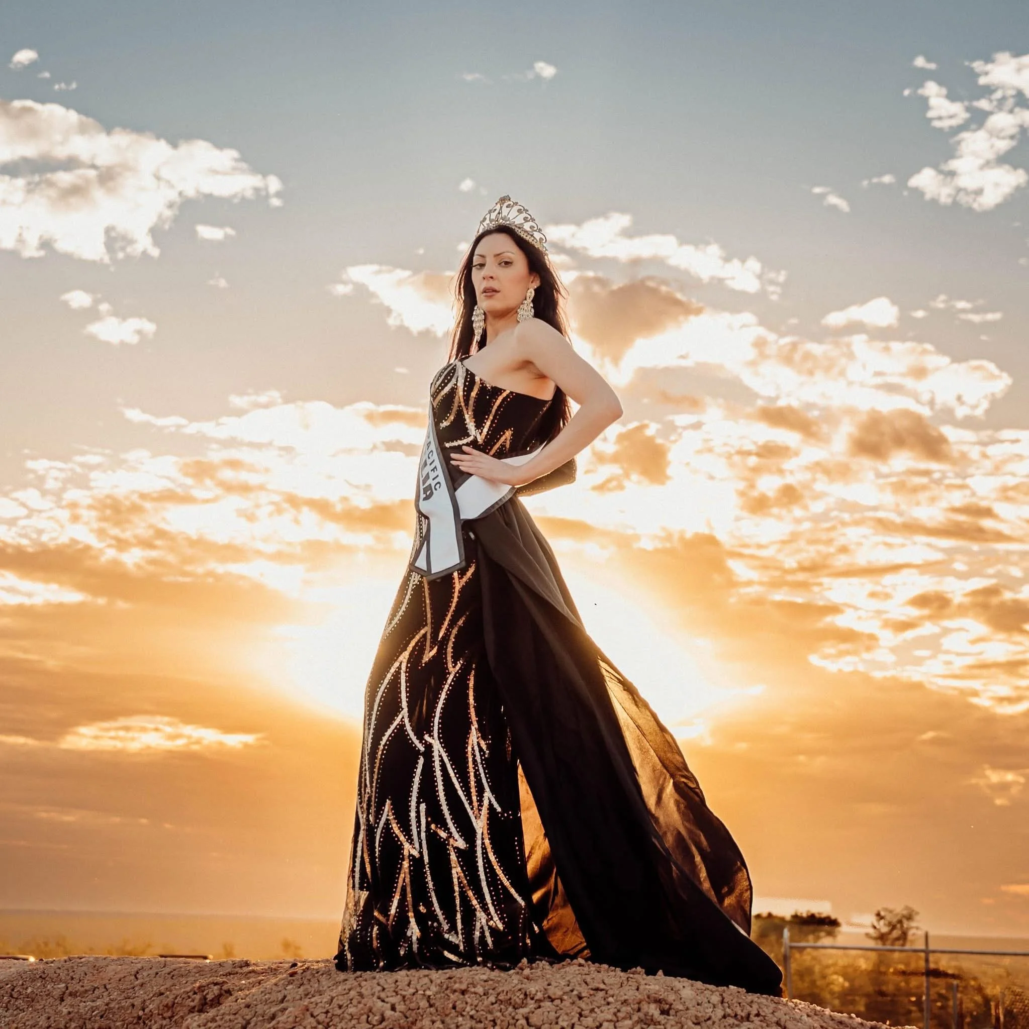 Woman wearing a crown and a black dress with gold patterns stands outdoors during sunset, with a dramatic sky and clouds in the background.