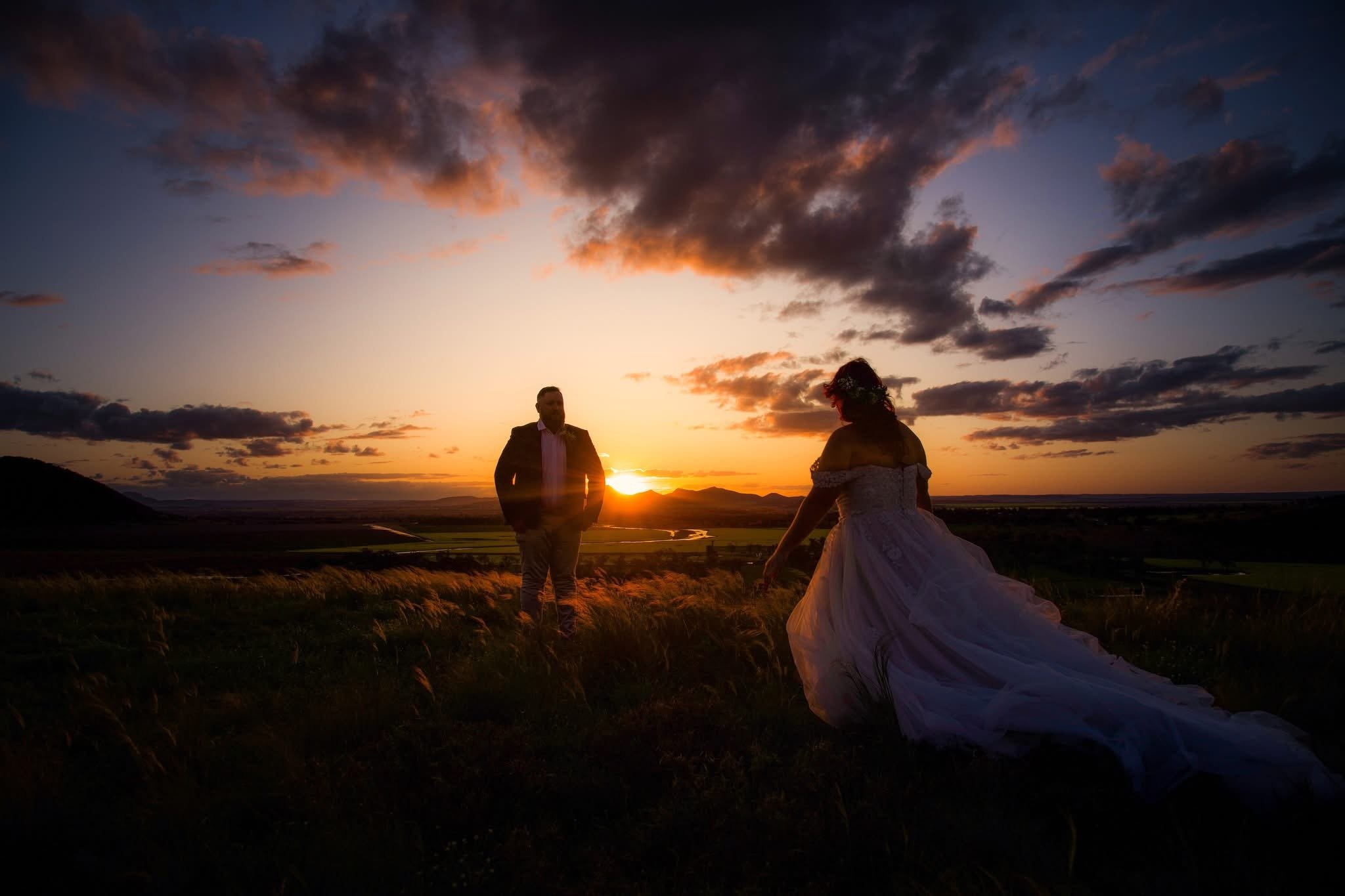 A couple, dressed in formal attire, stands in a grassy field at sunset; the man faces the woman, who is wearing a flowing gown, with the sunset behind them and scattered clouds in the sky.
