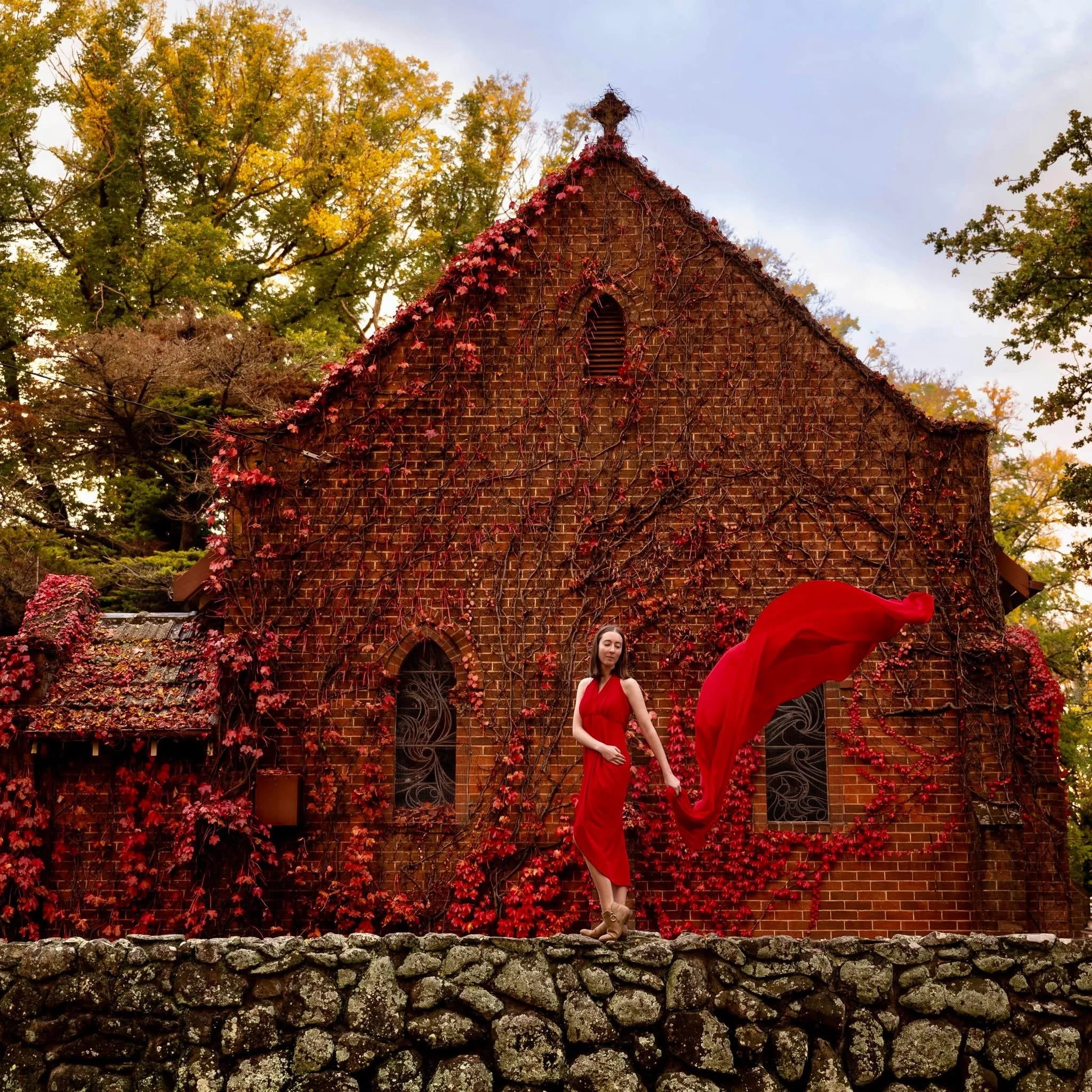 A woman in a red dress standing on a stone wall in front of a brick building covered with ivy and red leaves. She is holding a flowing red piece of fabric.