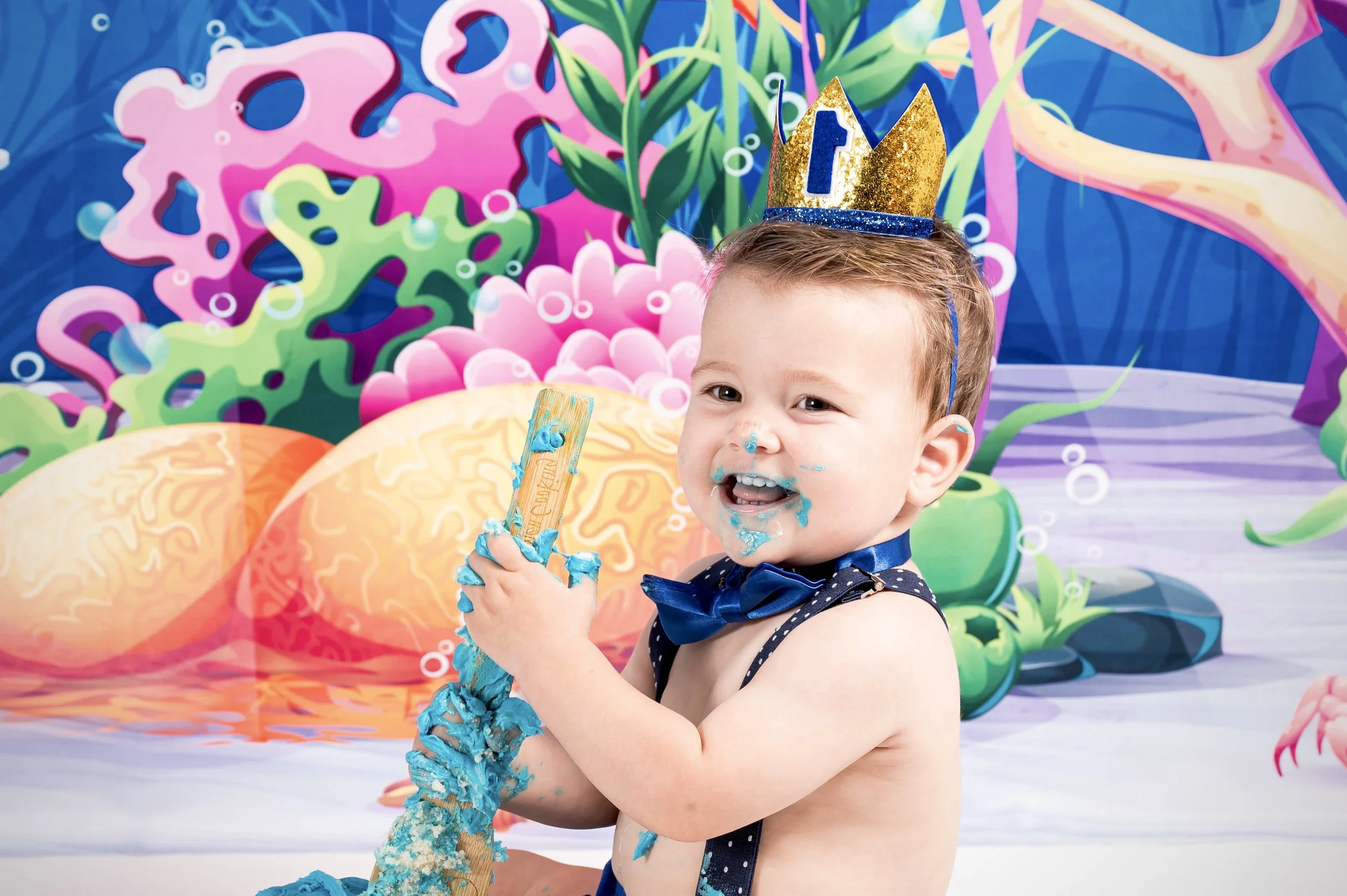 A young boy celebrating his birthday, wearing a blue bow tie and a glittery gold and blue crown, holding a party horn covered in blue frosting. The background features a colorful, underwater-themed mural with coral, seaweed, and shells.