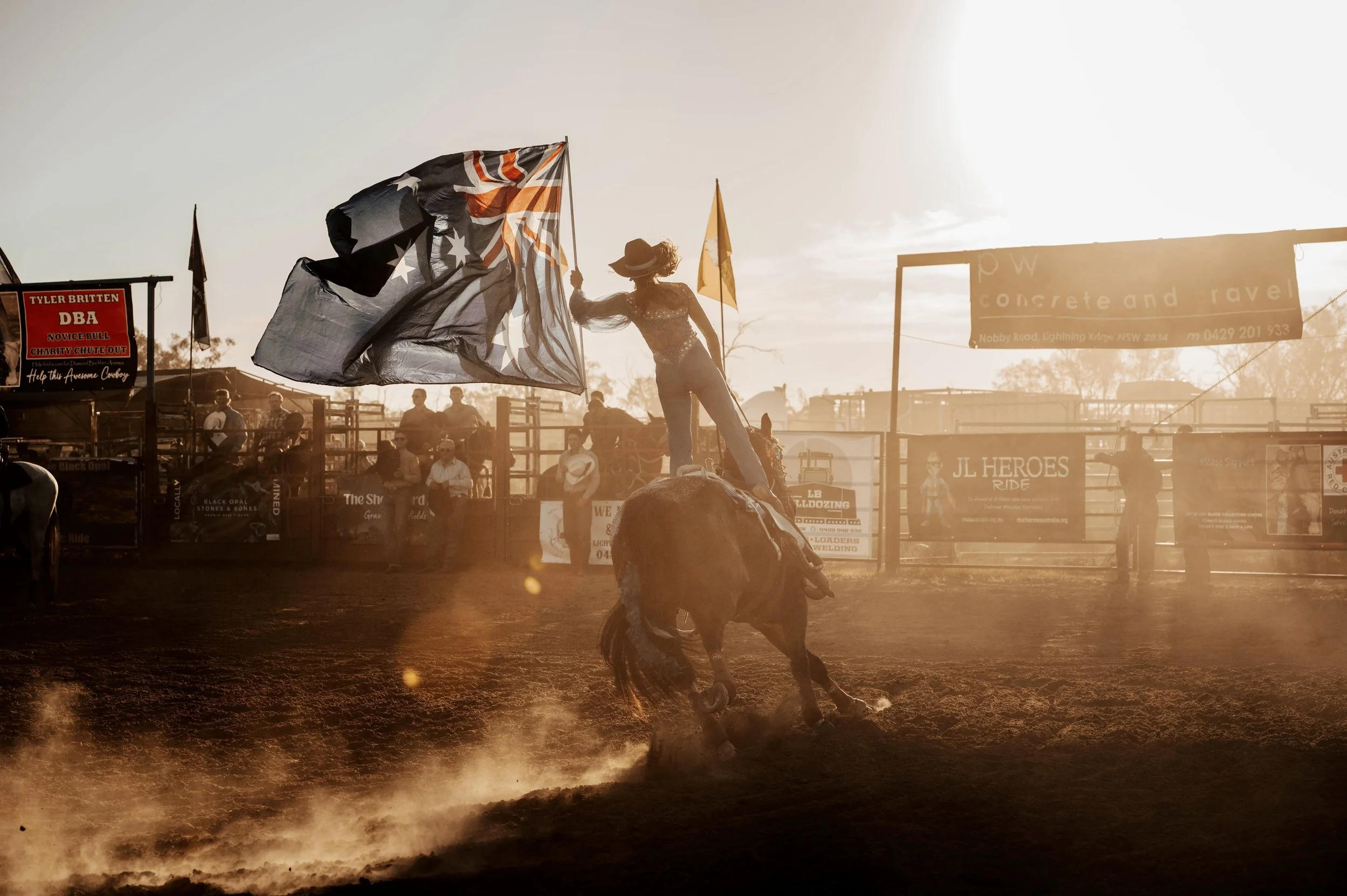 A cowgirl performing a rodeo trick on a bucking horse during sunset at a rodeo arena, holding a large flag with a black, orange, and white design, with spectators watching in the background.