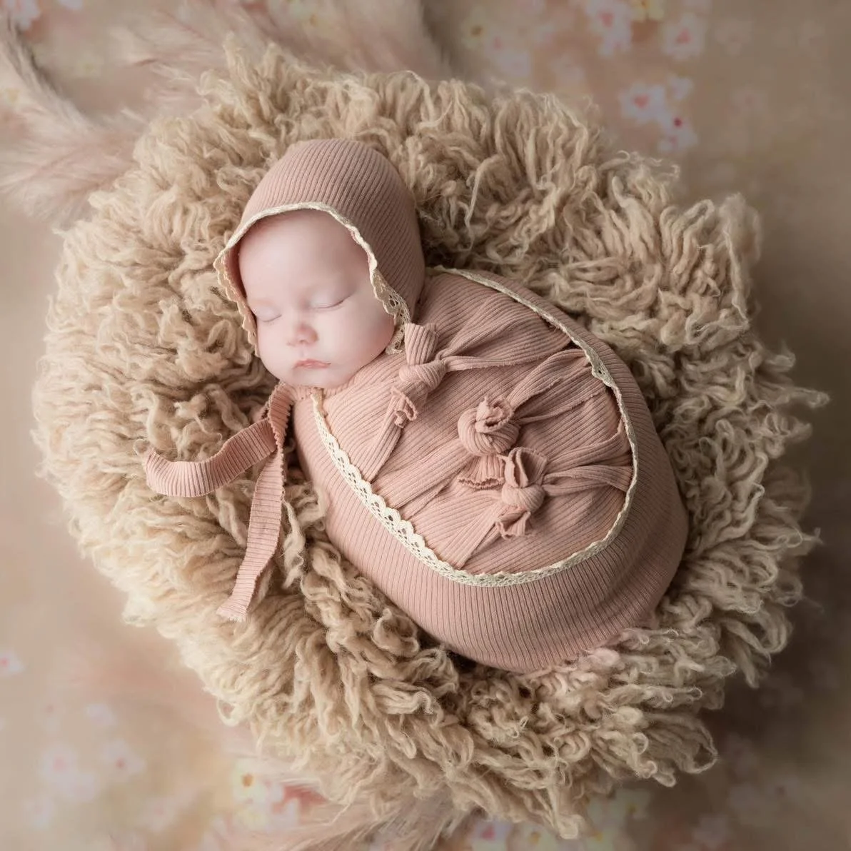 A sleeping baby swaddled in a pink outfit with a matching bonnet, lying on a fluffy beige blanket.