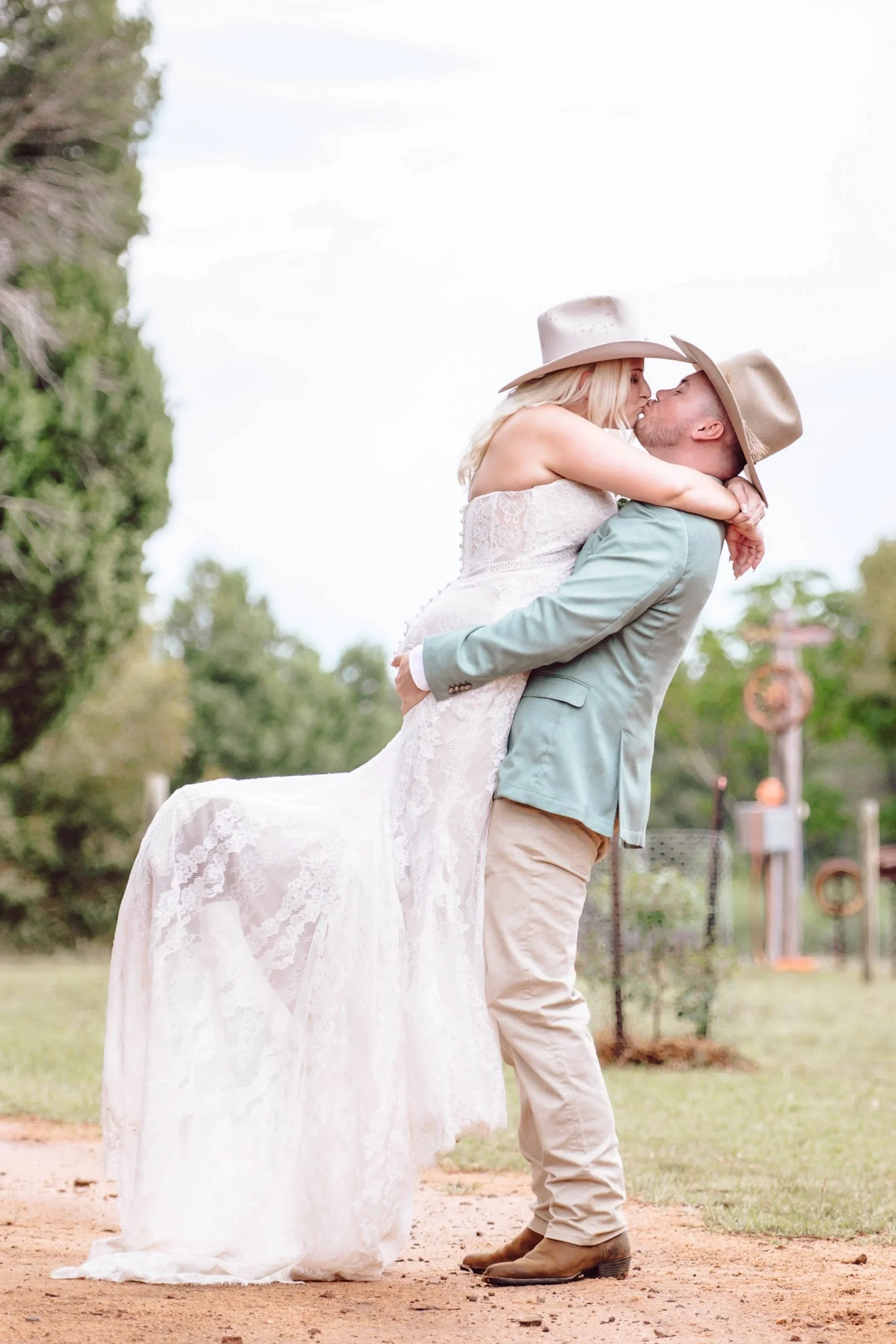 A couple sharing a kiss outdoors; the man is lifting the woman, who is wearing a white lace dress, and both are wearing cowboy hats.