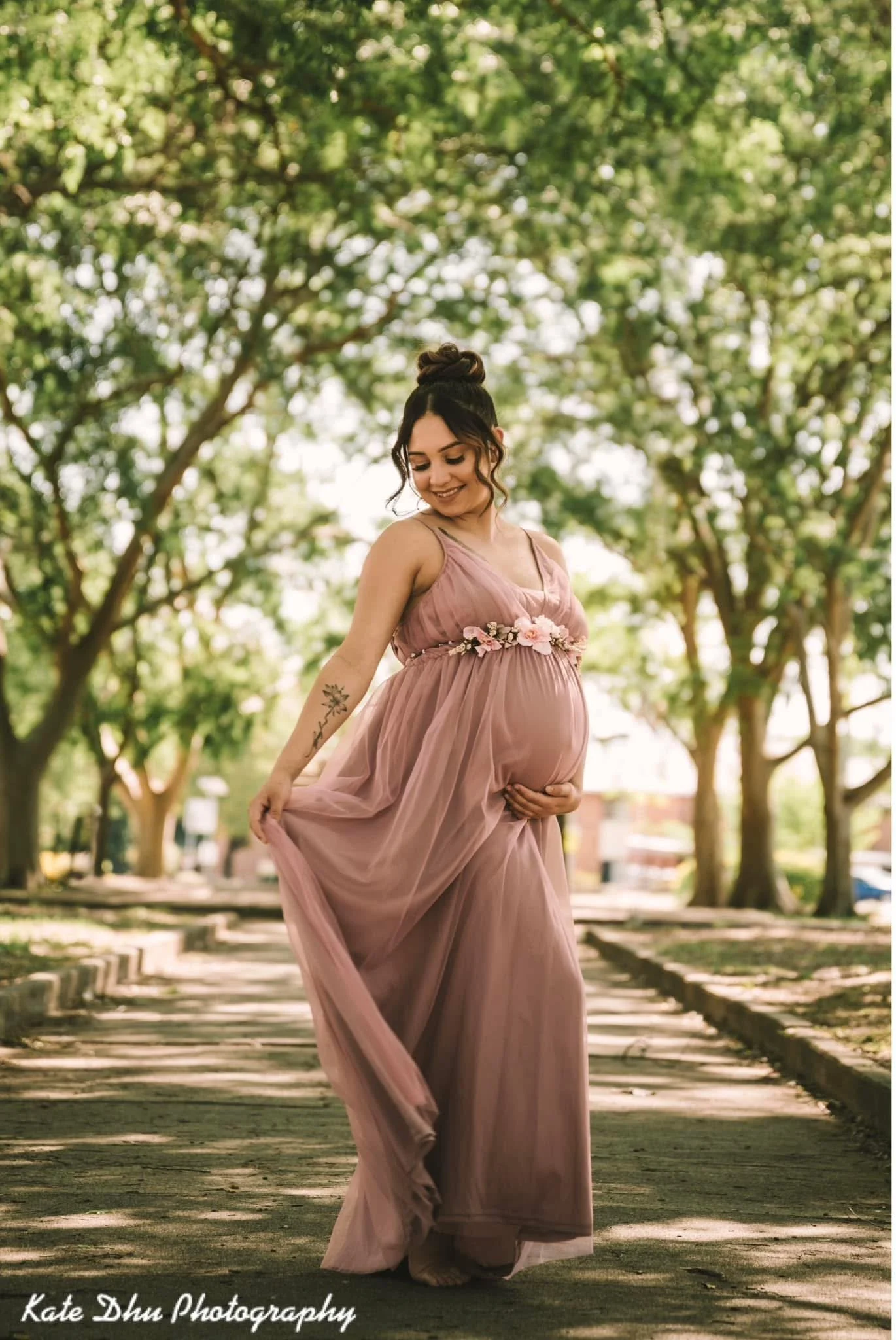Pregnant woman in pink flowing dress with floral belt, standing on a park path surrounded by green trees, looking down and smiling.