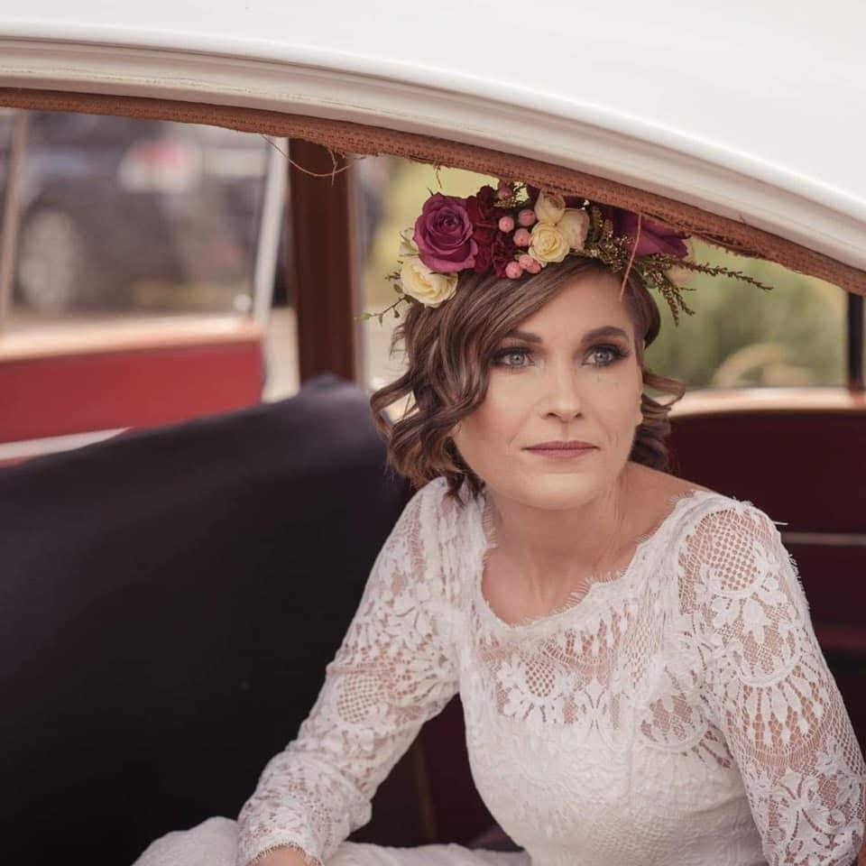 A woman with short brown hair styled in loose curls wearing a white lace top and a floral crown with purple and yellow flowers sitting in a vintage car.