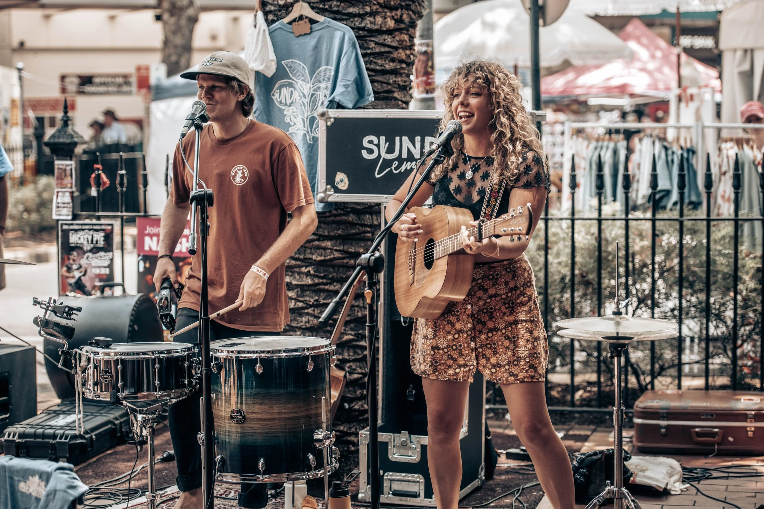 A man and woman perform music outdoors, woman playing guitar and singing, man playing drums, with a sign reading 'SUND' behind them at a street fair.