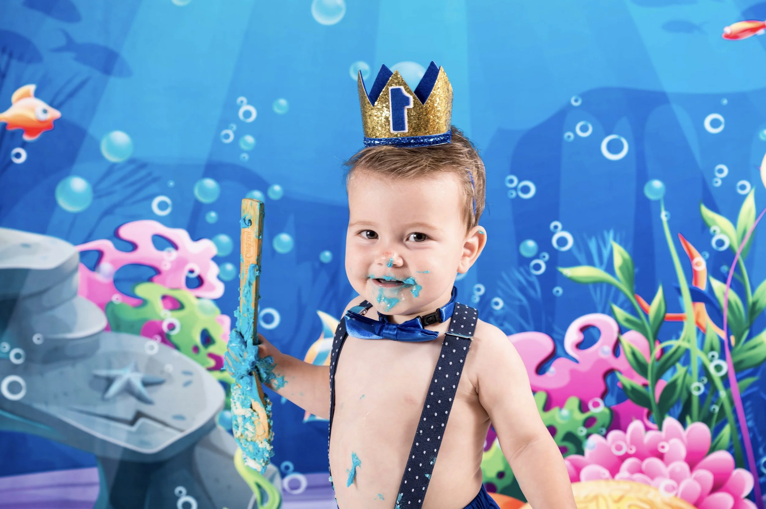 Young boy at birthday party with blue sea-themed backdrop, wearing black suspenders and a blue polka-dotted bow tie, gold and blue crown, messy with blue cake around mouth and on shirt, holding a cake knife.
