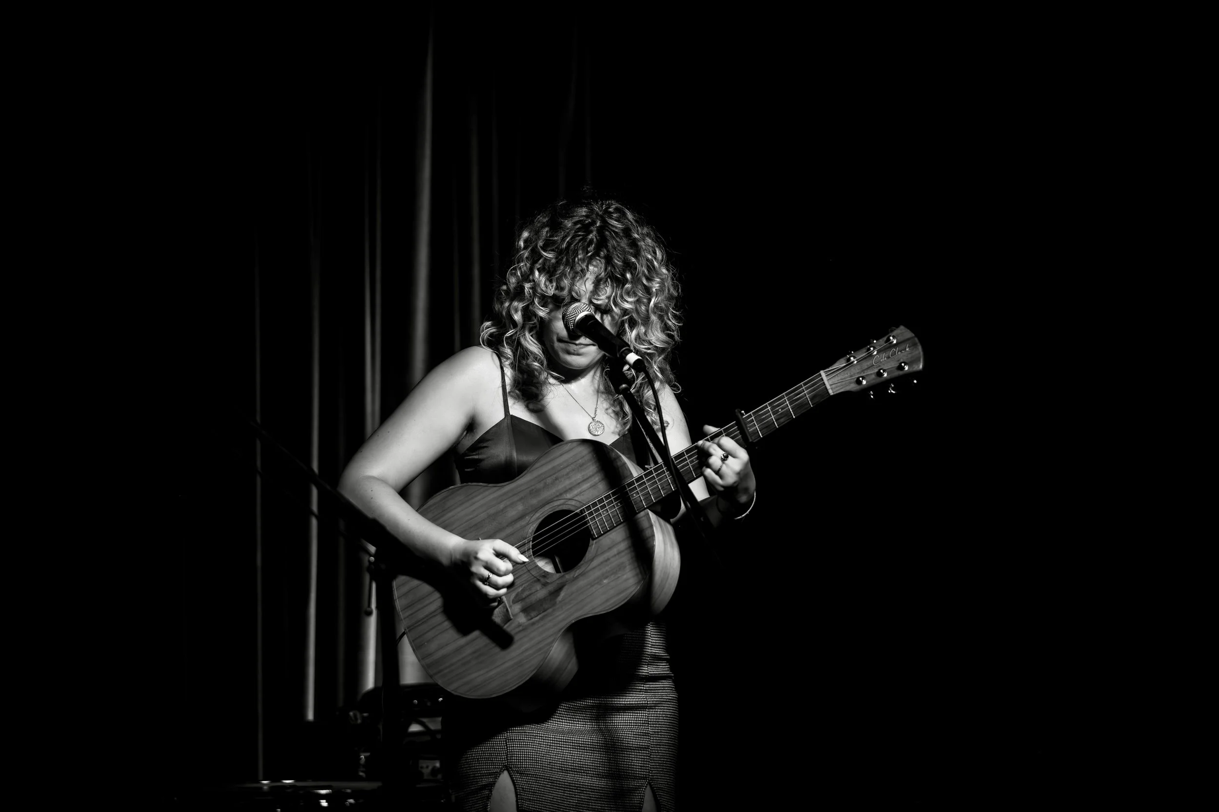 A female musician with curly hair playing an acoustic guitar on stage with a microphone in front of her, in black and white.
