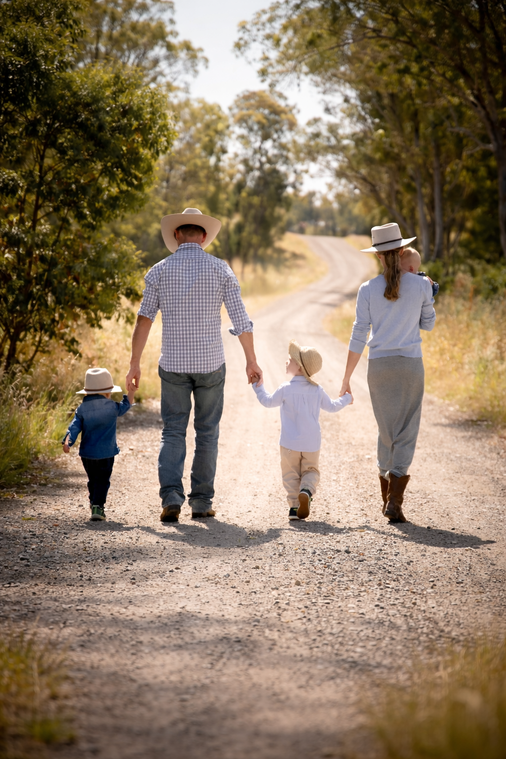 Family of four walking on a dirt road surrounded by trees; two adults and two children, all wearing hats, holding hands, with one child carrying a baby.