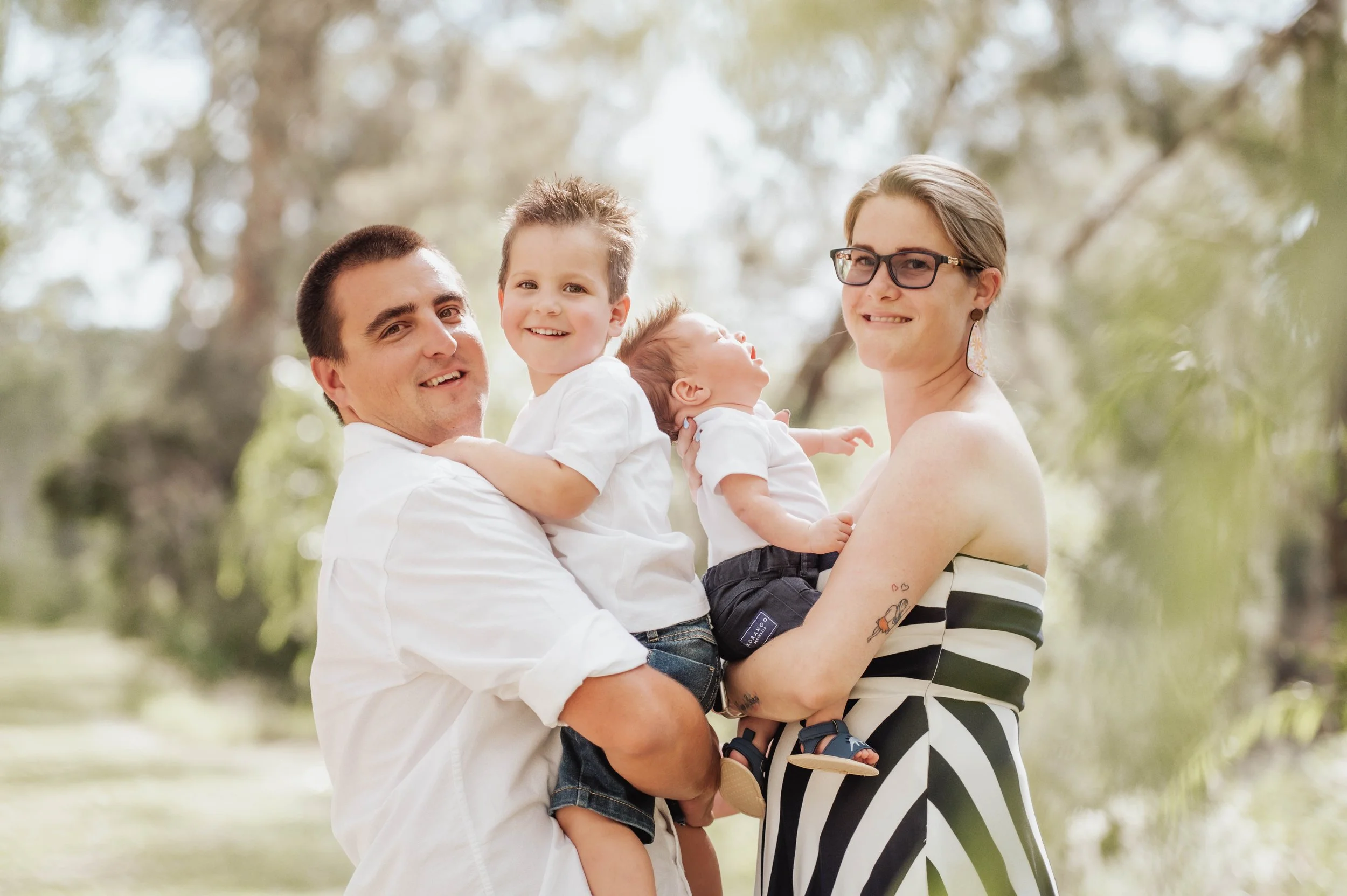 Family of four outdoors, with a man holding a young boy, a woman holding a baby, and trees in the background.