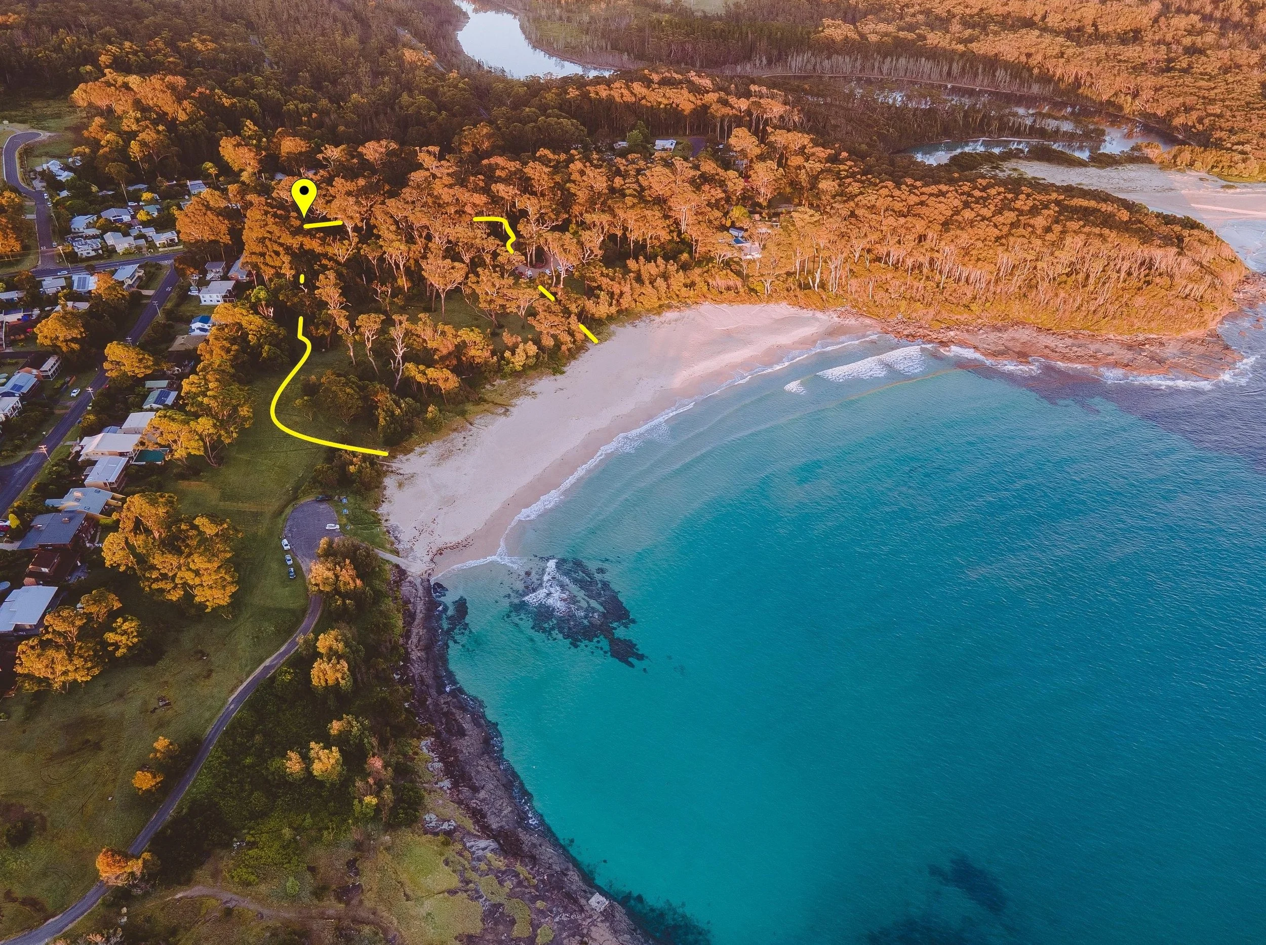 Aerial view of a coastal area with a sandy beach, blue ocean water, dense forest, residential houses, and winding roads.