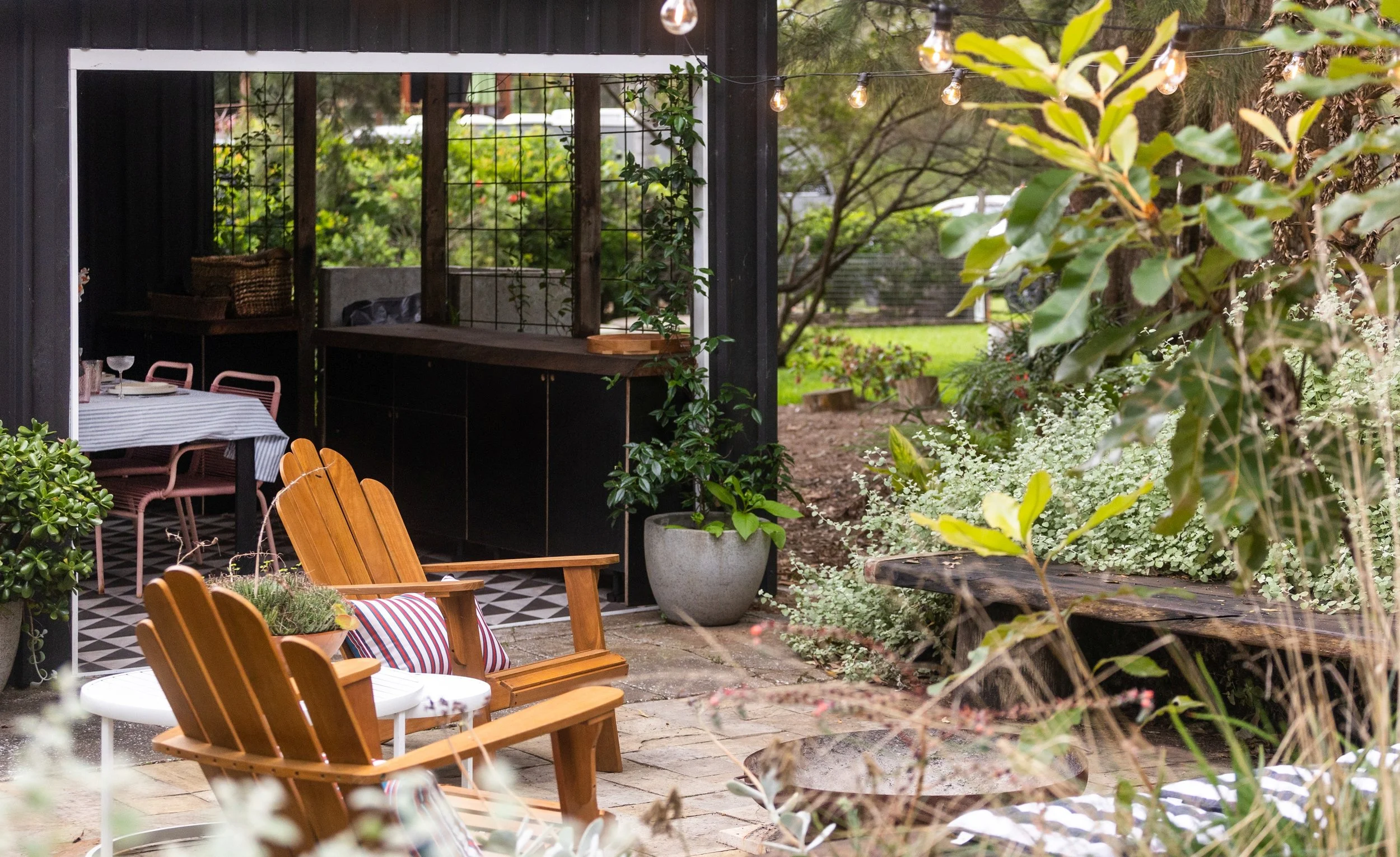 Overgrown outdoor patio with wooden Adirondack chairs, potted plants, and string lights under trees.