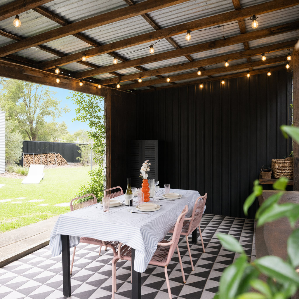 Outdoor dining area with a white-striped tablecloth, set for six, under a wooden pergola with string lights, overlooking a grassy backyard with trees and stacked firewood.