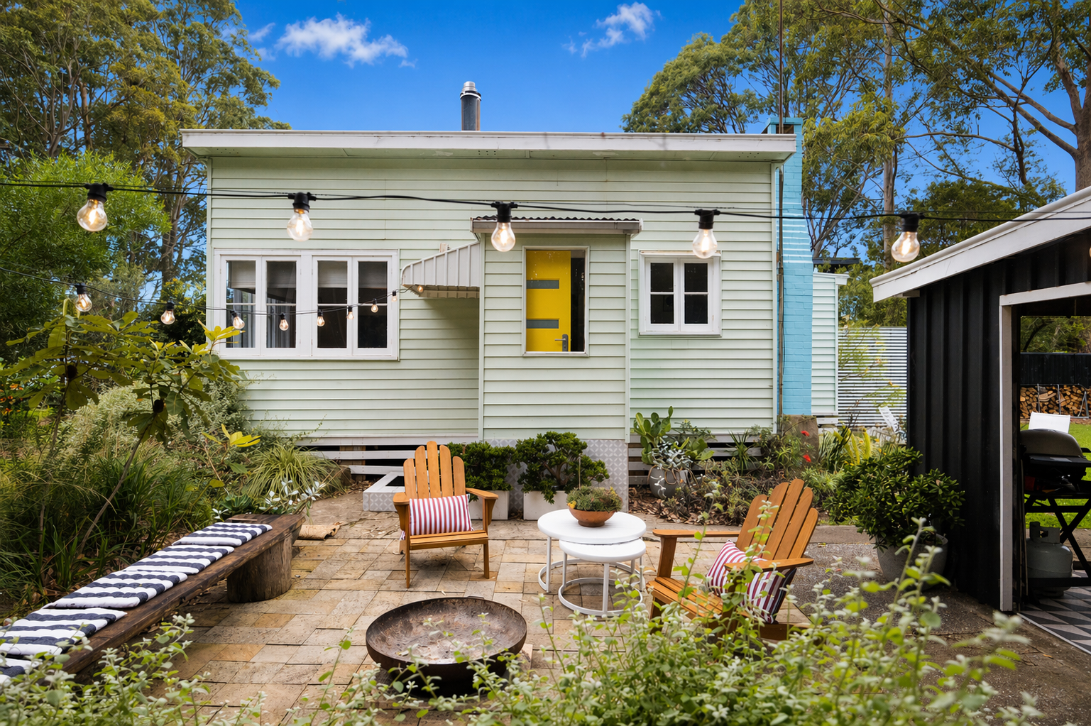 Backyard patio with string lights, garden, wooden and metal chairs, tables, and firepit in front of a pastel green house with yellow door.