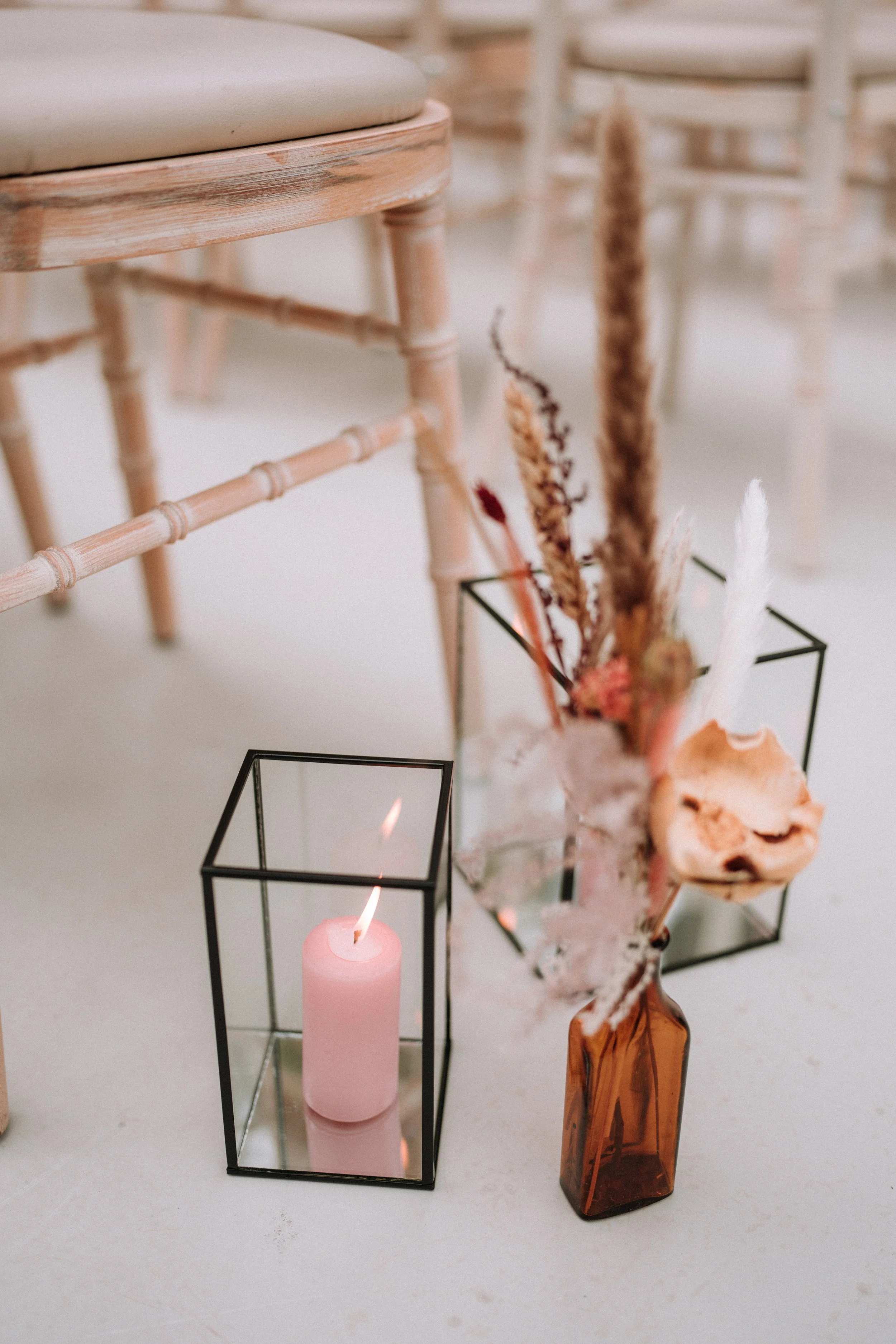 A pink candle in a black metal square holder, a brown glass vase with dried flowers and ornamental branches, and a piece of coral on a light-colored surface.
