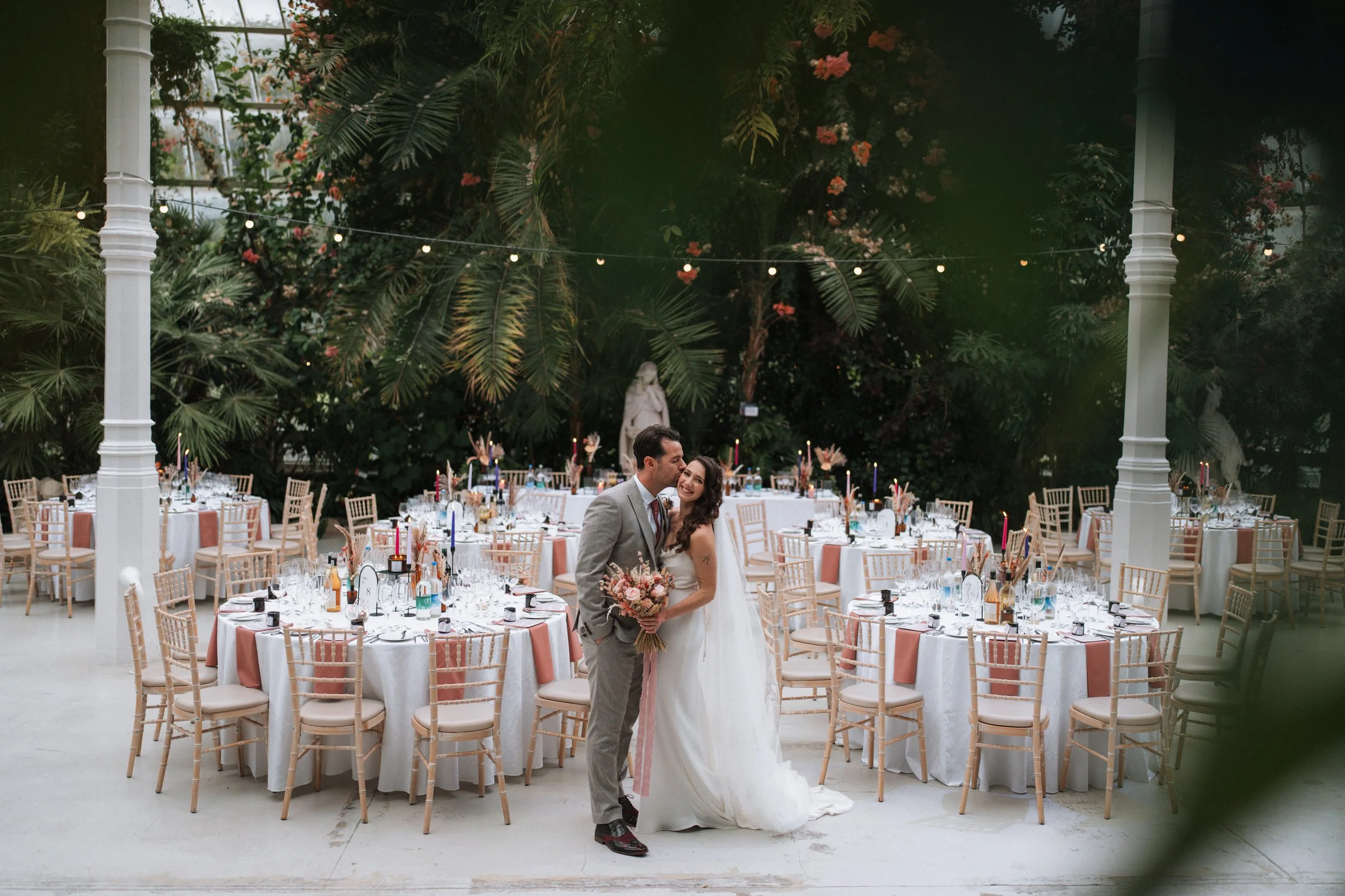 A wedding reception in an indoor garden with decorated tables and a bride and groom embracing in front of them.