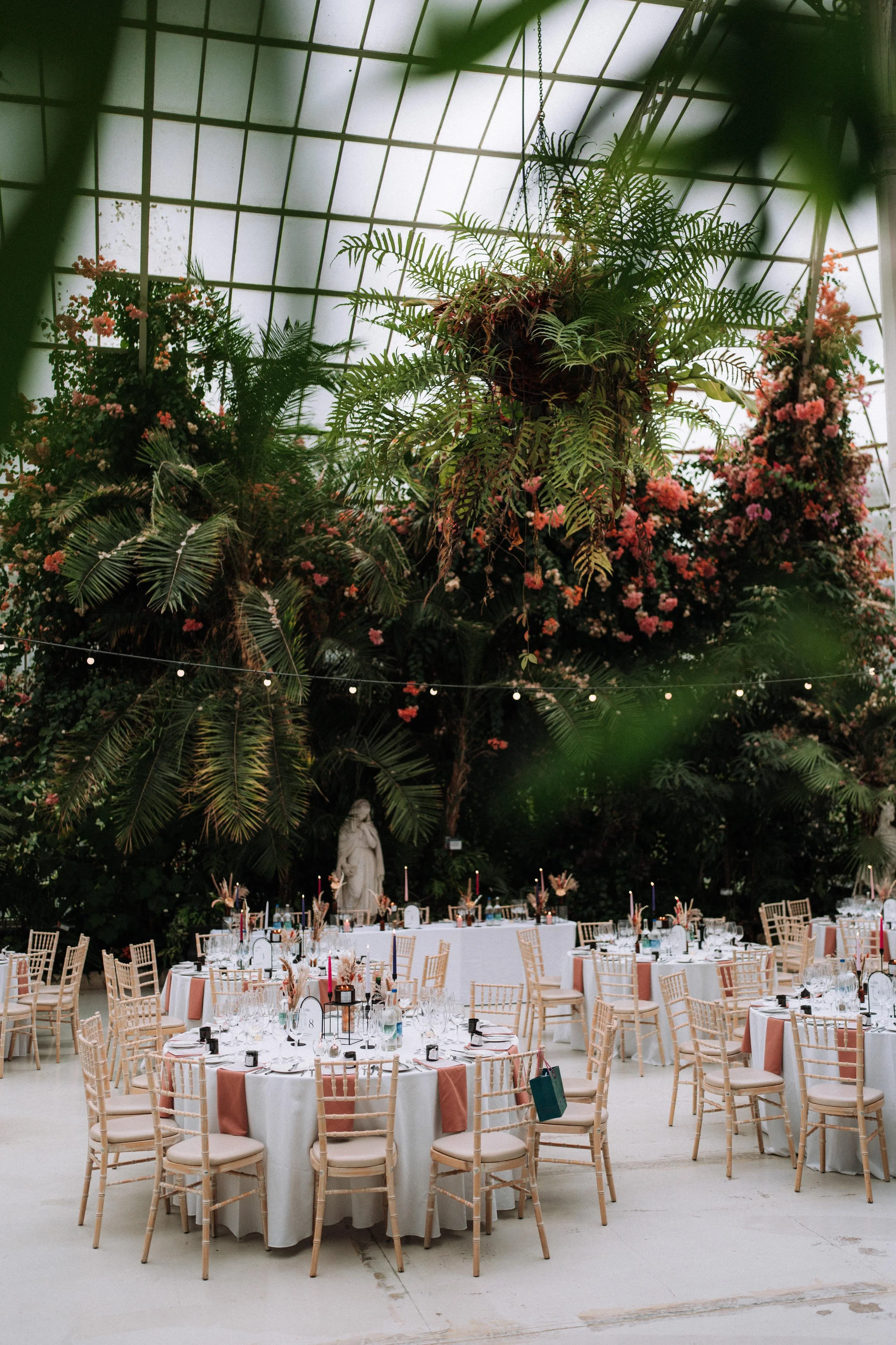 Indoor event space decorated with round tables covered with white tablecloths and pink table runners, surrounded by wooden chairs with soft cushions. Each table is set with glassware, silverware, and floral centerpieces featuring pink and neutral tones. A large statue of a woman stands at the back, flanked by lush green trees and flowering bushes, with string lights hanging overhead in a greenhouse-style ceiling.